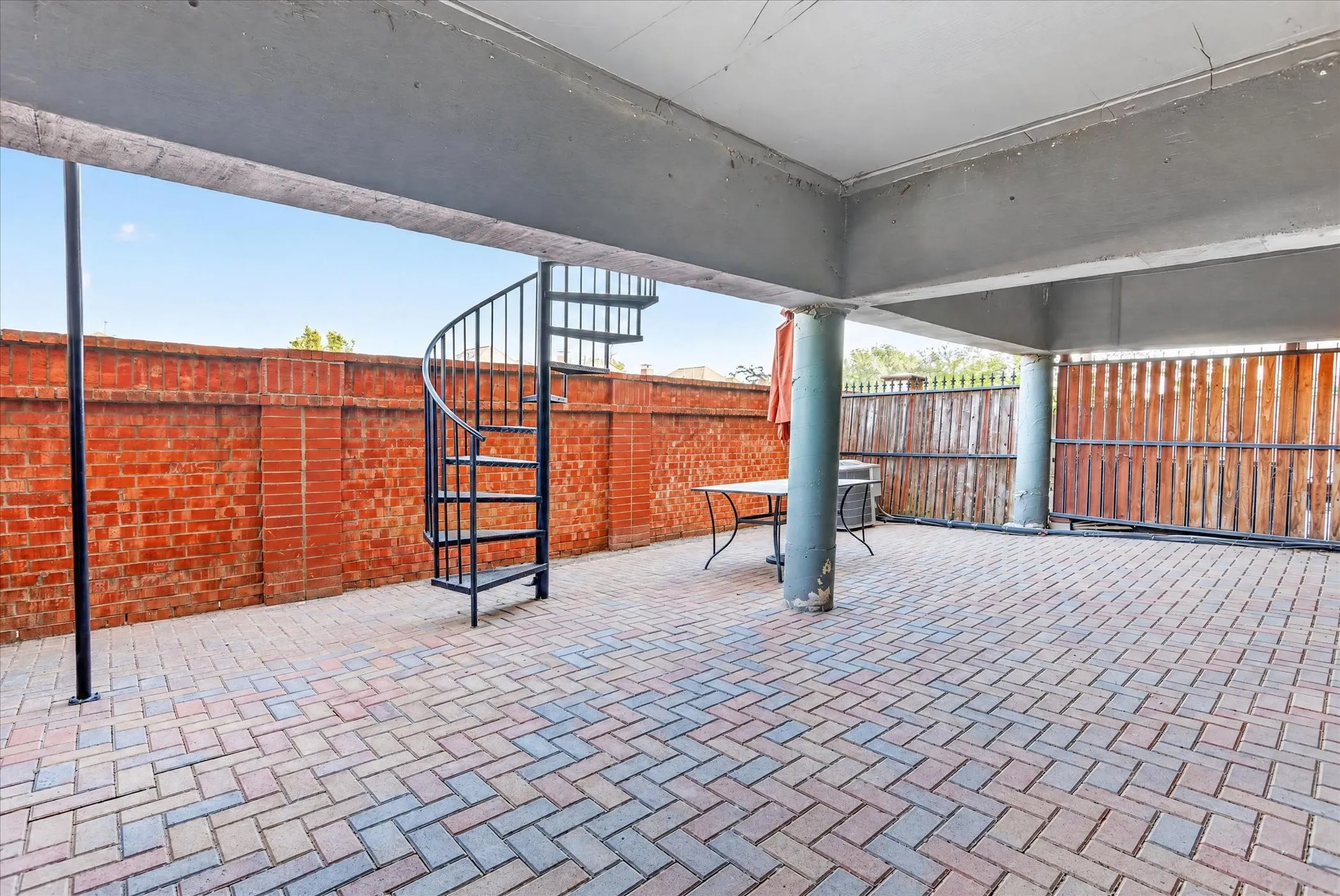 Covered patio featuring herringbone-patterned brick pavers, a wrought-iron spiral staircase, structural concrete pillars, and a perimeter masonry wall