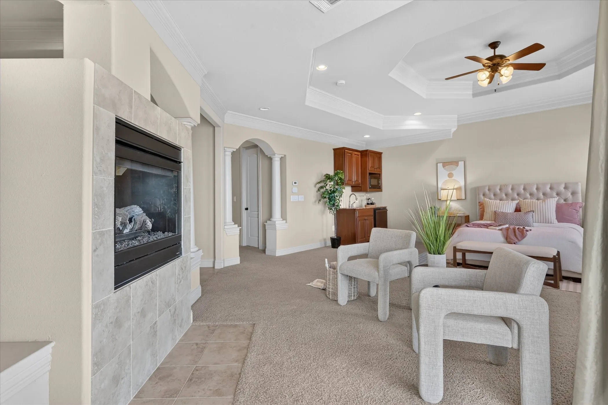 Tray ceiling with recessed lighting, fireplace with stone-finish surround, built-in cabinetry with integrated sink, and architectural column detailing