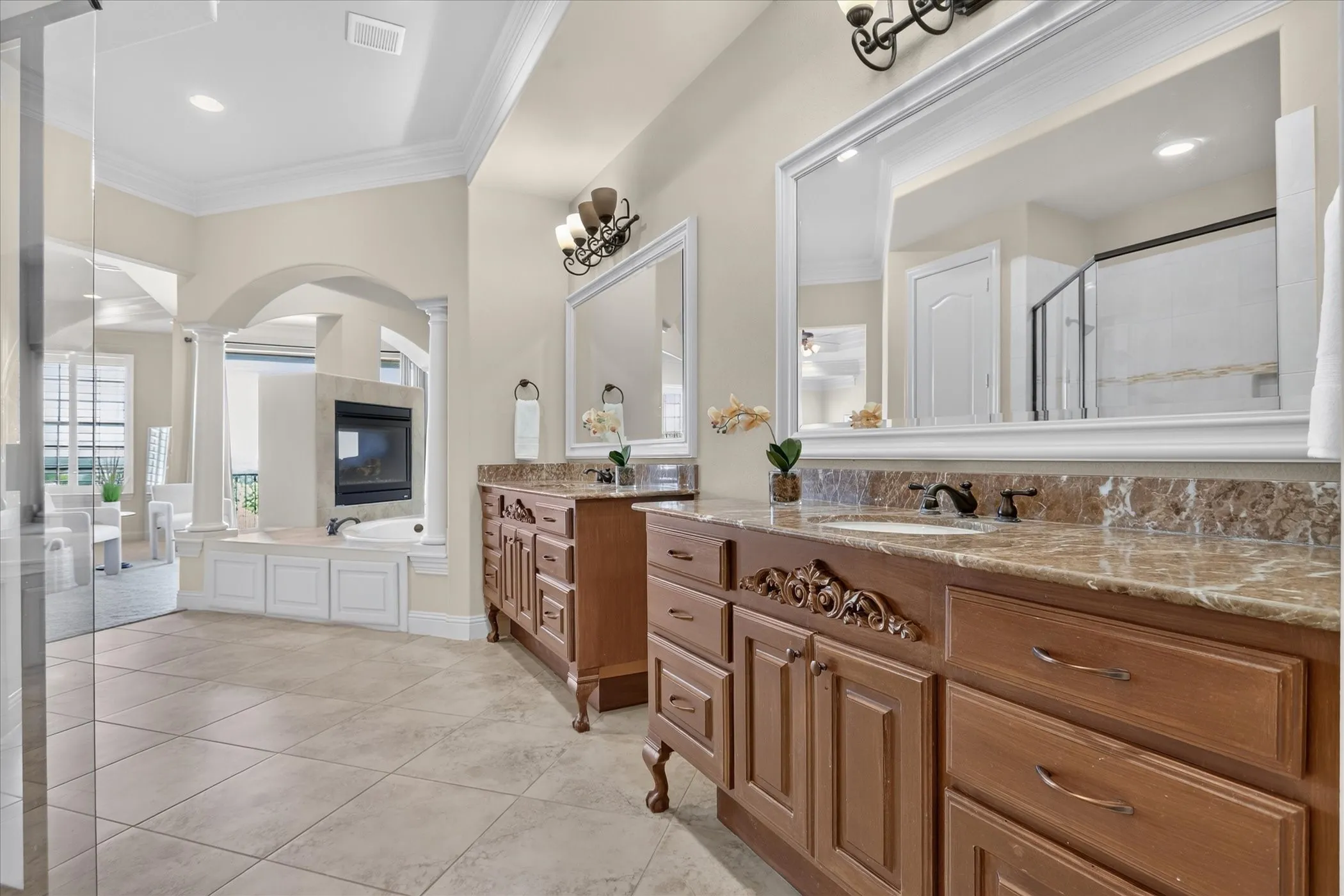 Dual wood vanities featuring stone countertops, large framed wall mirrors, a deep soaking tub, a built-in fireplace, and tile flooring
