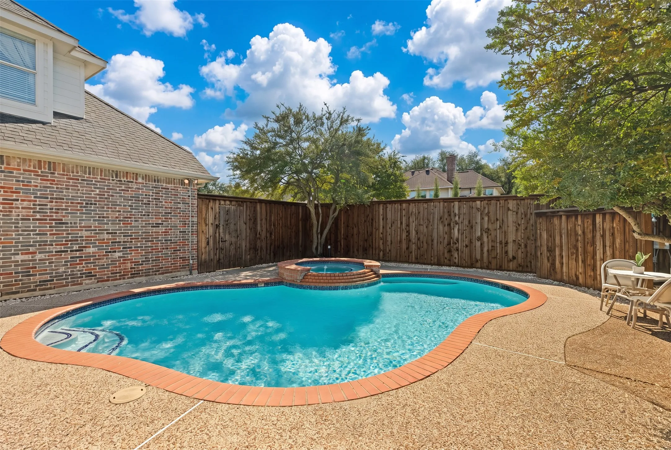 In-ground swimming pool with an integrated elevated spa, surrounded by textured aggregate decking, brick masonry exterior, and a tall wood privacy fence