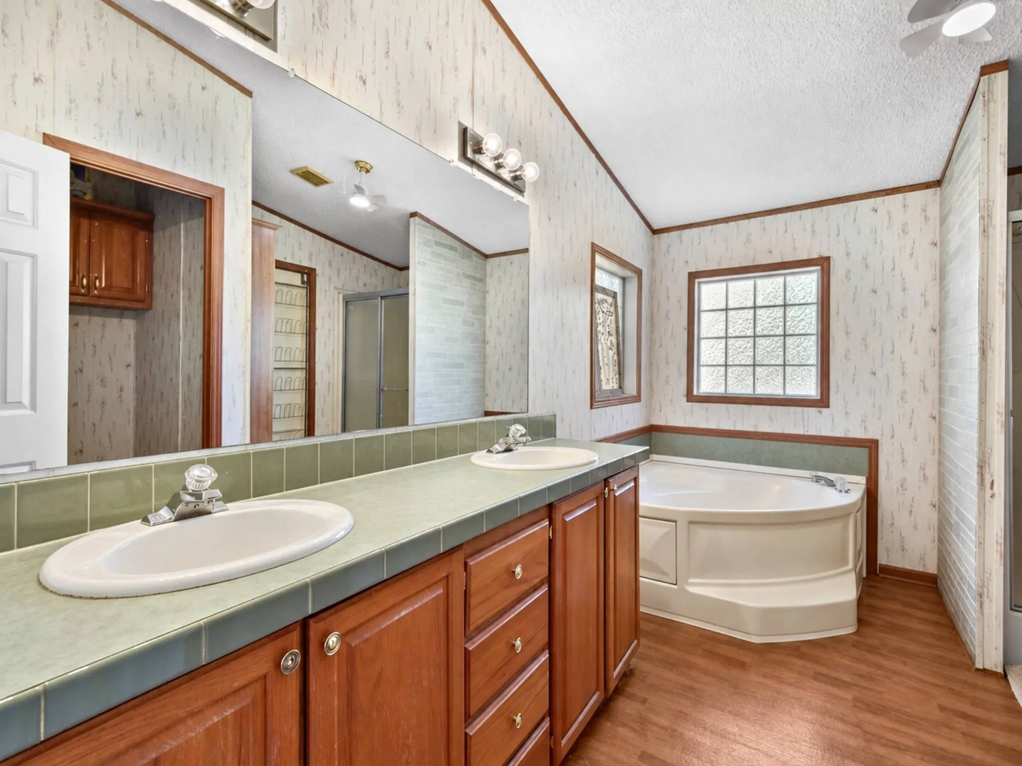 Dual-sink vanity with tile countertop, wood-finish cabinetry, corner soaking tub, and block-pattern privacy window
