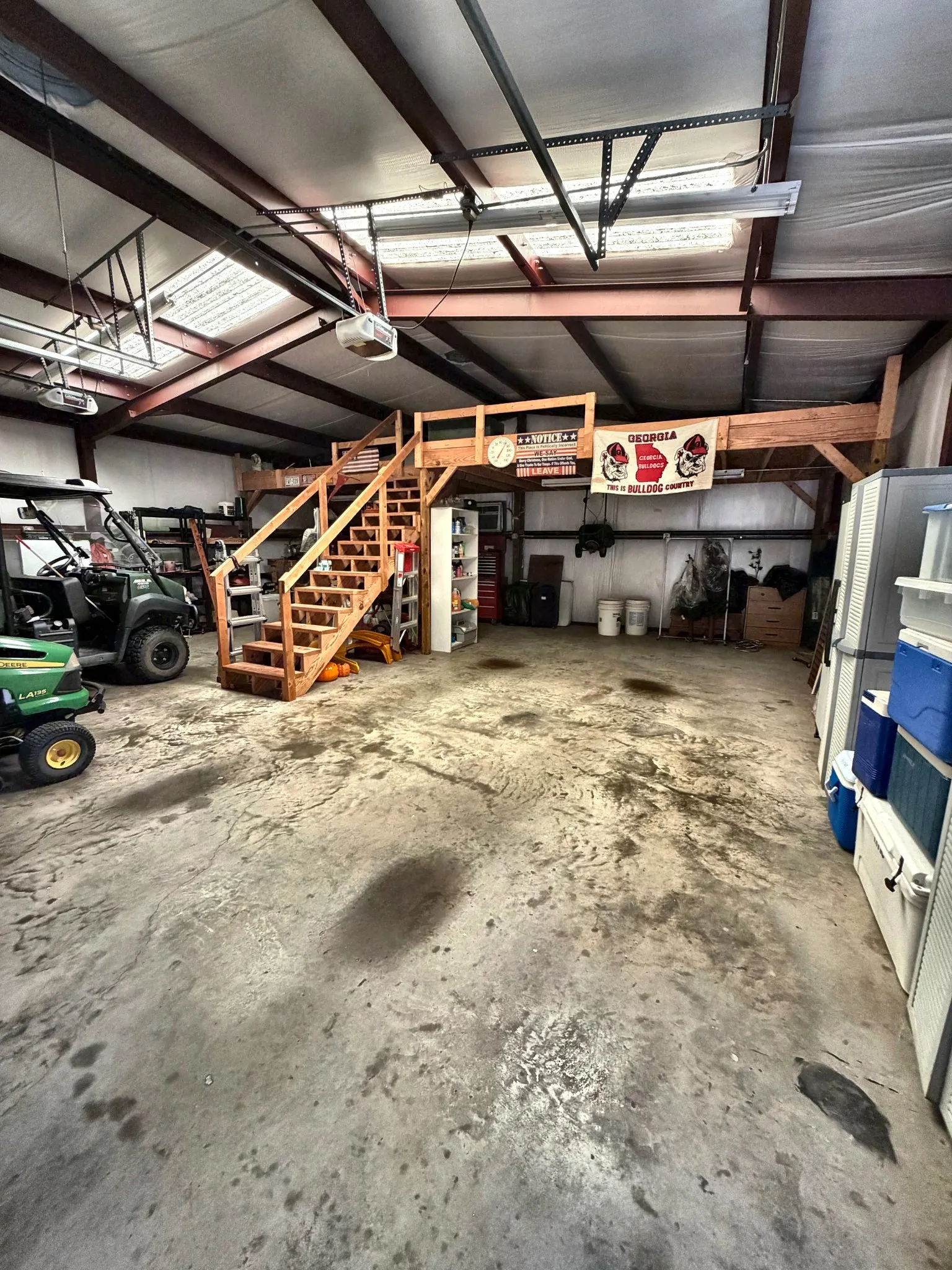 Spacious workshop featuring a concrete floor, exposed steel beams, translucent roof panels, and an elevated wooden loft with a built-in staircase