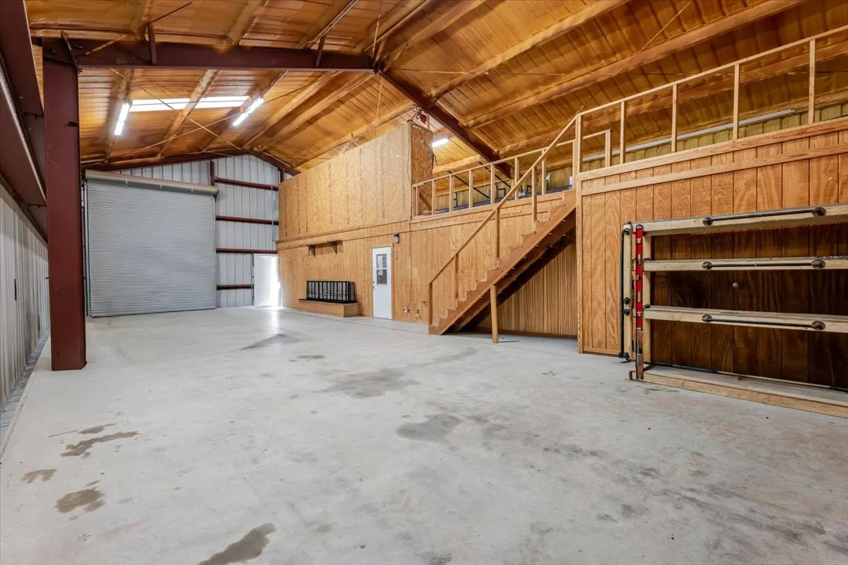 Spacious Warehouse featuring a concrete floor, high vaulted wood-paneled ceiling, roll-up overhead door, built-in wooden shelving, and an integrated elevated loft area with a wood staircase