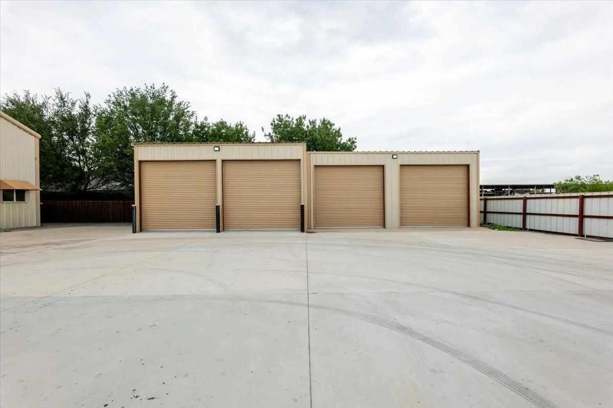 Detached four-car metal garage building featuring a wide concrete apron, roll-up doors, and exterior wall-mounted lighting
