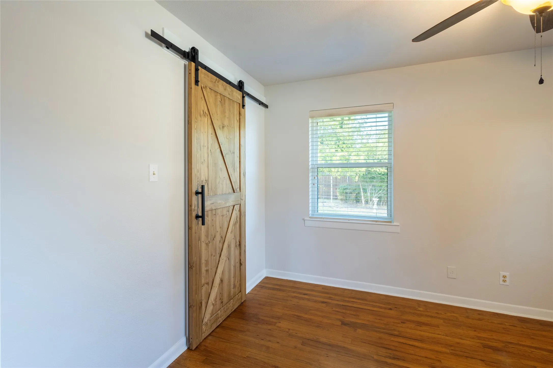 Wood-finish flooring, sliding barn door with matte black hardware, horizontal slat window blinds, and a ceiling fan