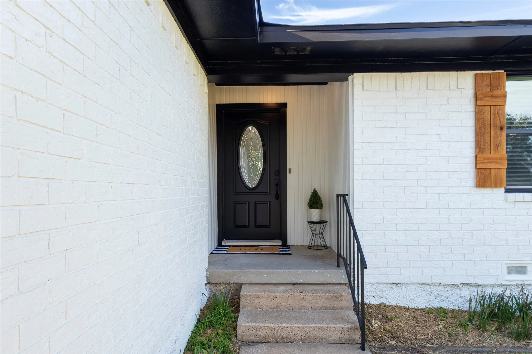 White brick exterior featuring a black front door with an oval glass insert, vertical wood-plank siding accents, natural wood shutters, and black metal railings