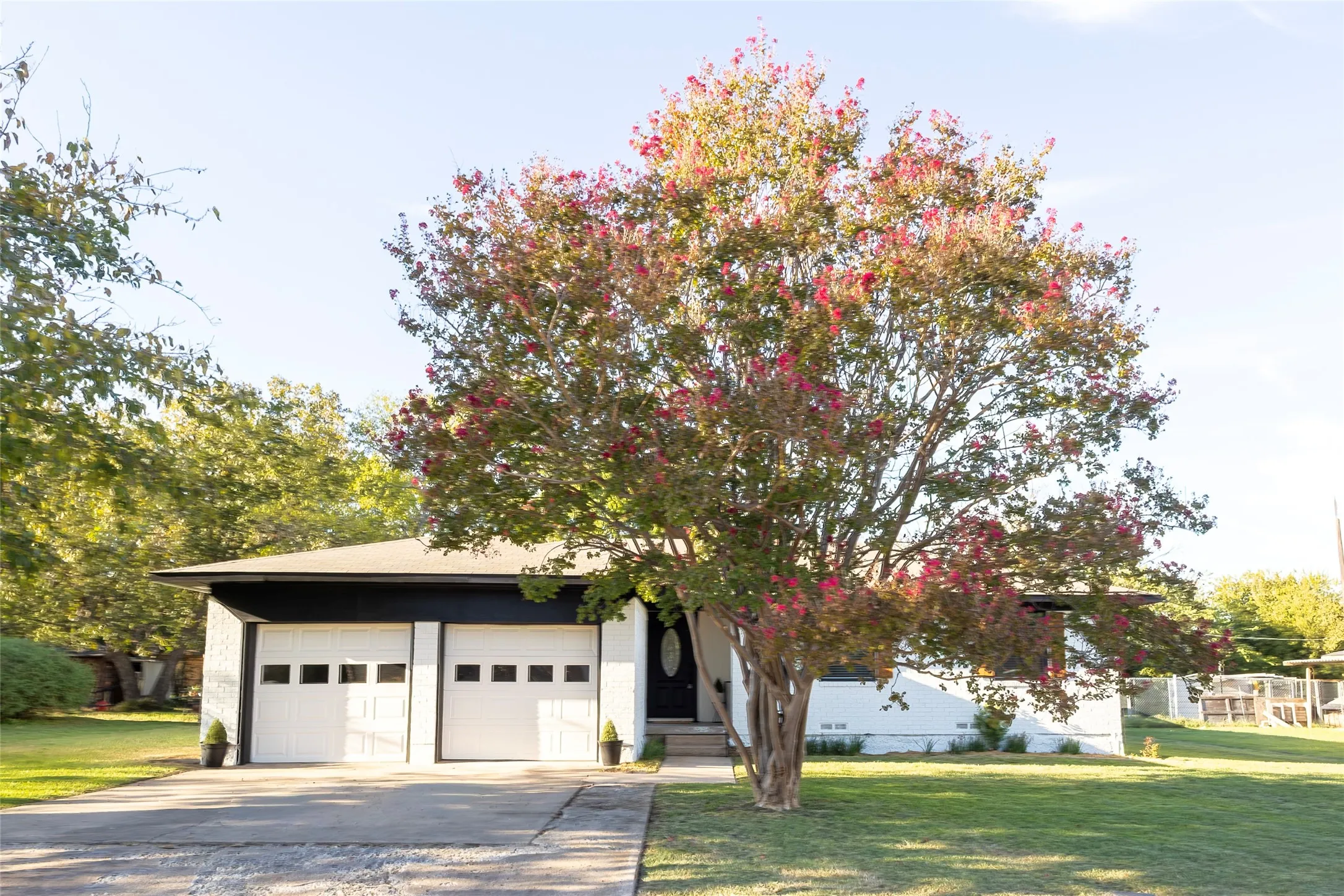Single-story exterior featuring a white-painted brick facade, two-car garage with rectangular window accents, concrete driveway, and mature landscaping