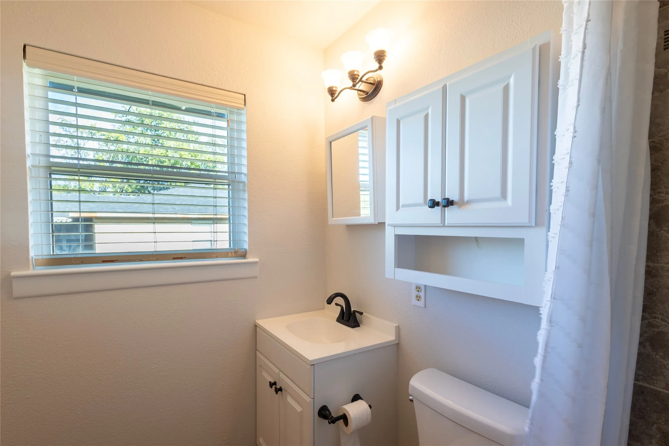 Bathroom featuring a vanity with an integrated sink, matte black faucet, wall-mounted white cabinetry, three-light fixture, and horizontal blind window treatment
