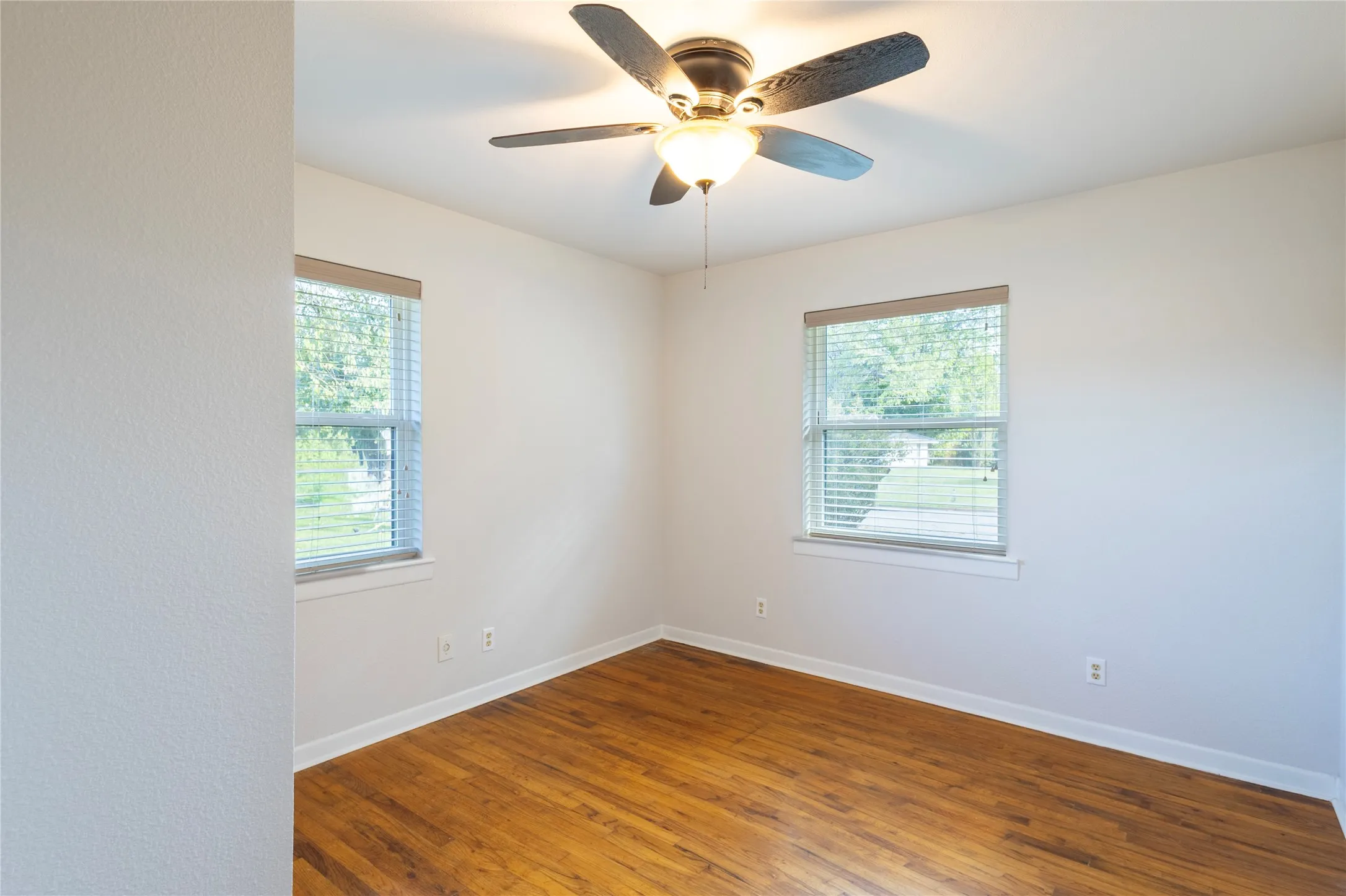 Room featuring wood-finish flooring, dual windows with blinds, and a central ceiling fan with integrated lighting