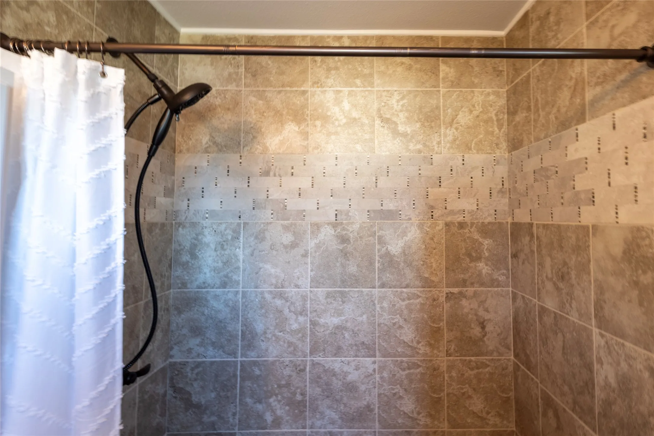 Shower surround featuring large-format neutral-toned tile, a horizontal decorative mosaic tile accent band, and an adjustable-height handheld shower fixture with a dark finish