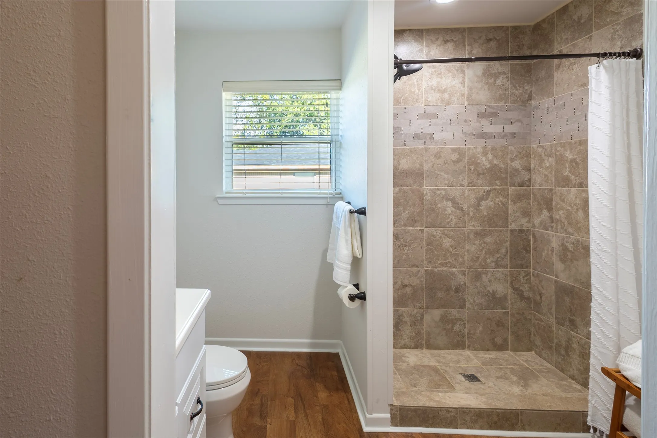 Walk-in shower with stone-tile surround, decorative mosaic tile inlay, wood-finish flooring, and window with horizontal blinds