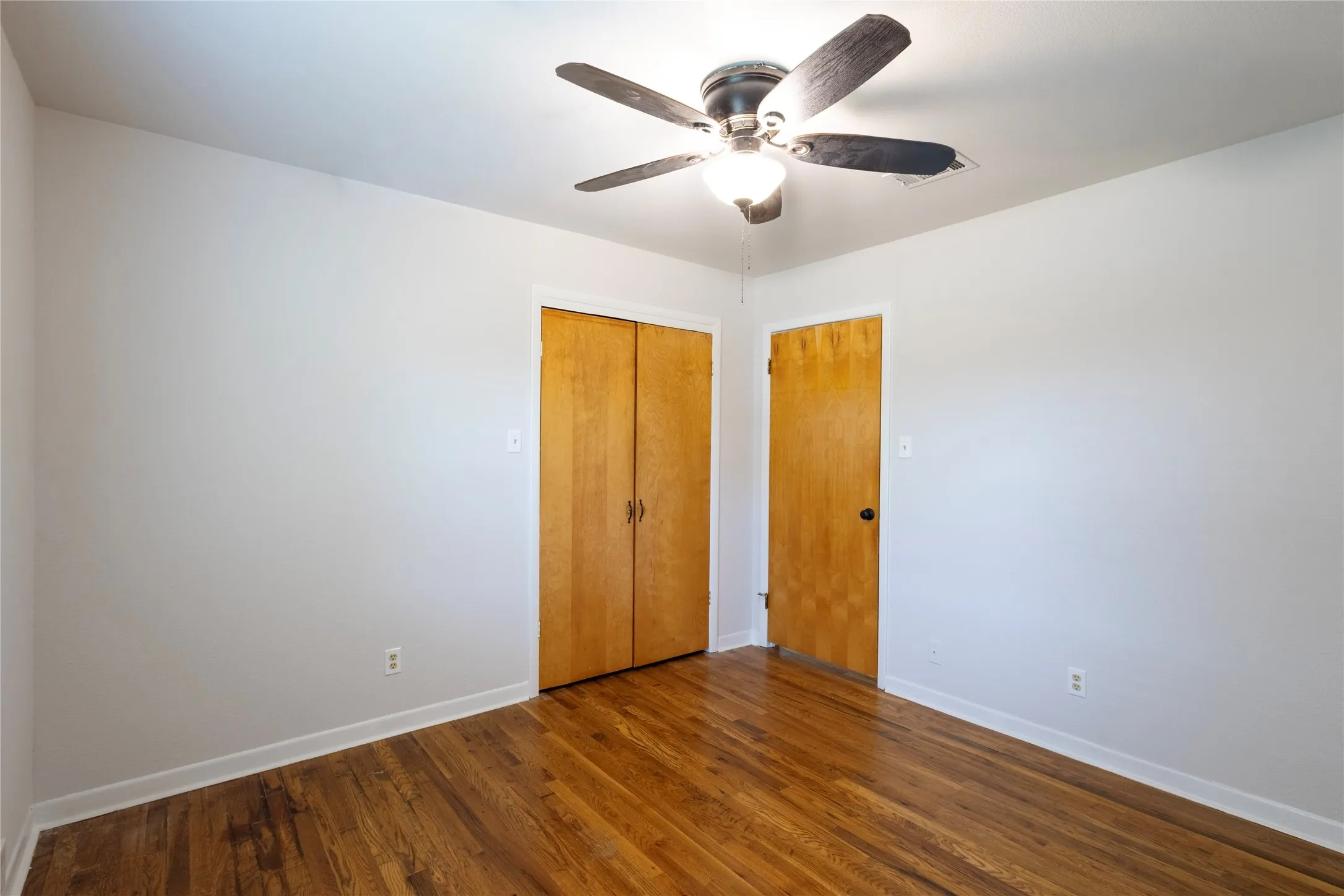 Room featuring wood-finish flooring, a central ceiling fan with integrated lighting, and natural wood-finish closet and passage doors