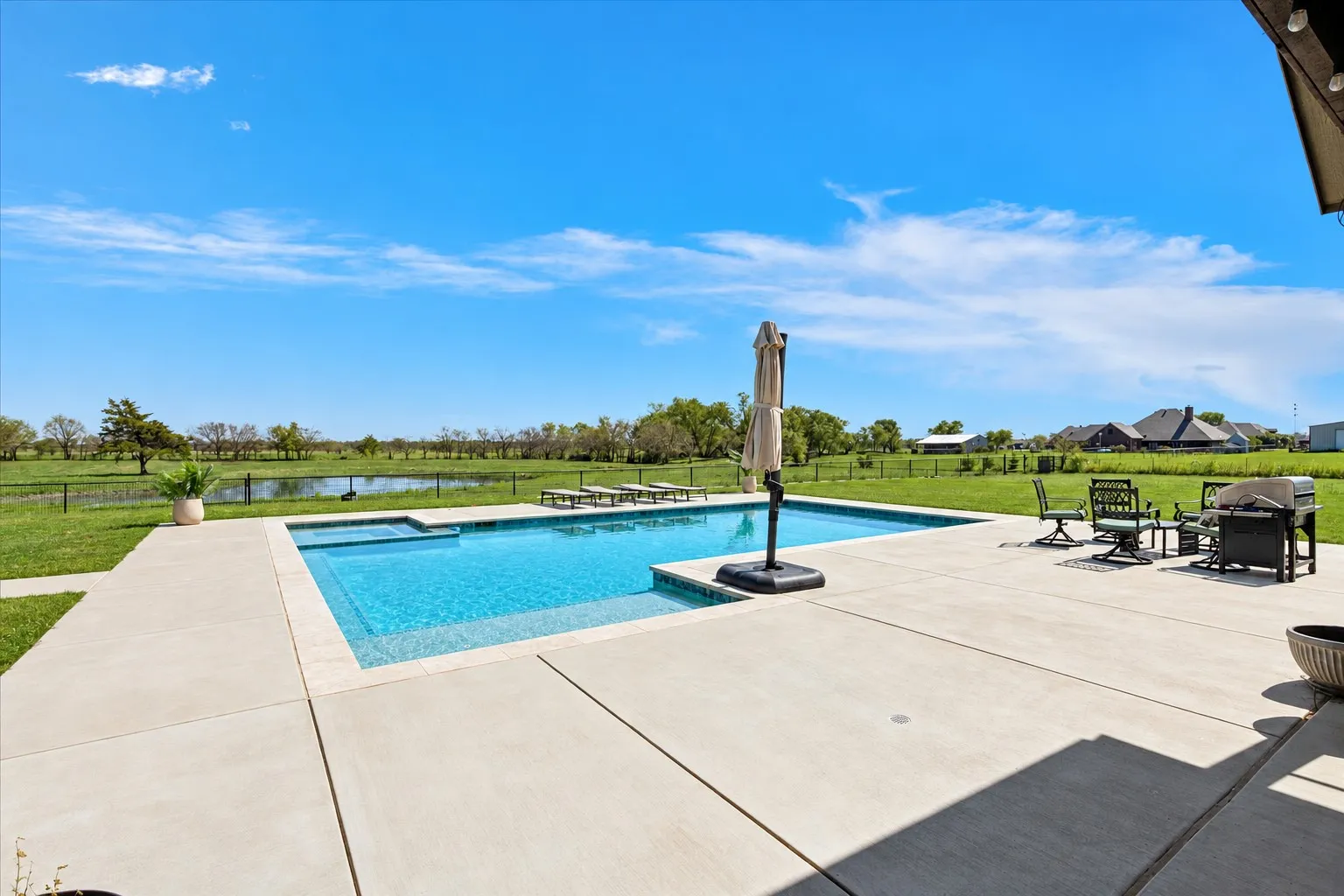 Inground swimming pool with tiled border and integrated spa, expansive concrete patio, and fenced grounds featuring a pond