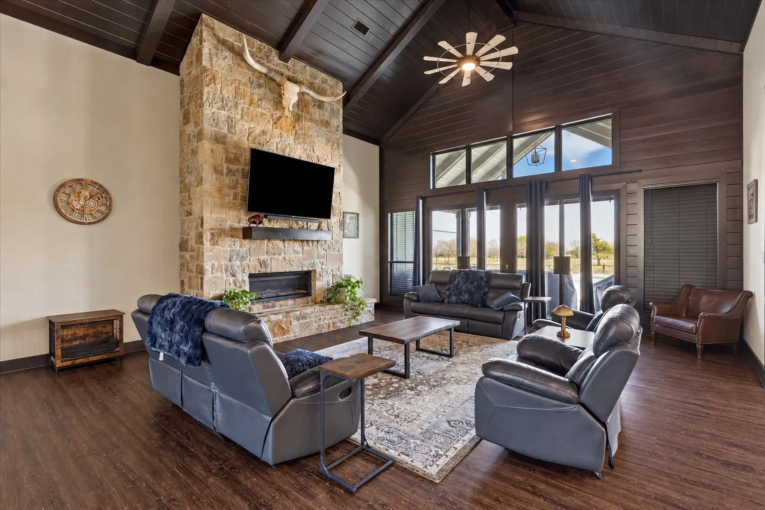 Vaulted ceiling with exposed timber beams, full-height stone fireplace, wood-finish flooring, and expansive transom windows