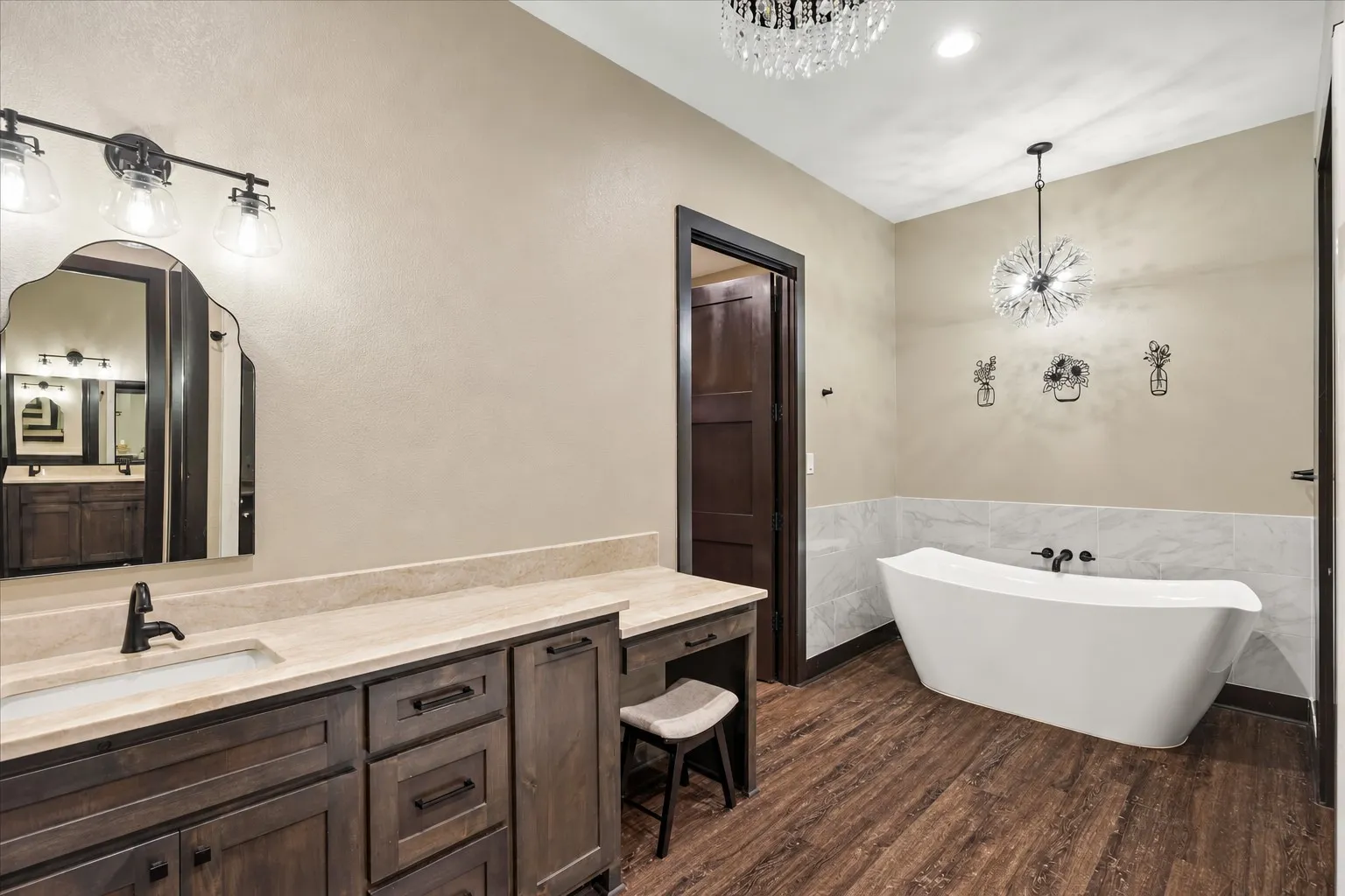 Freestanding soaking tub with a stone-finish wall surround, custom dark wood-finish cabinetry, stone countertop, matte black fixtures, and decorative pendant lighting