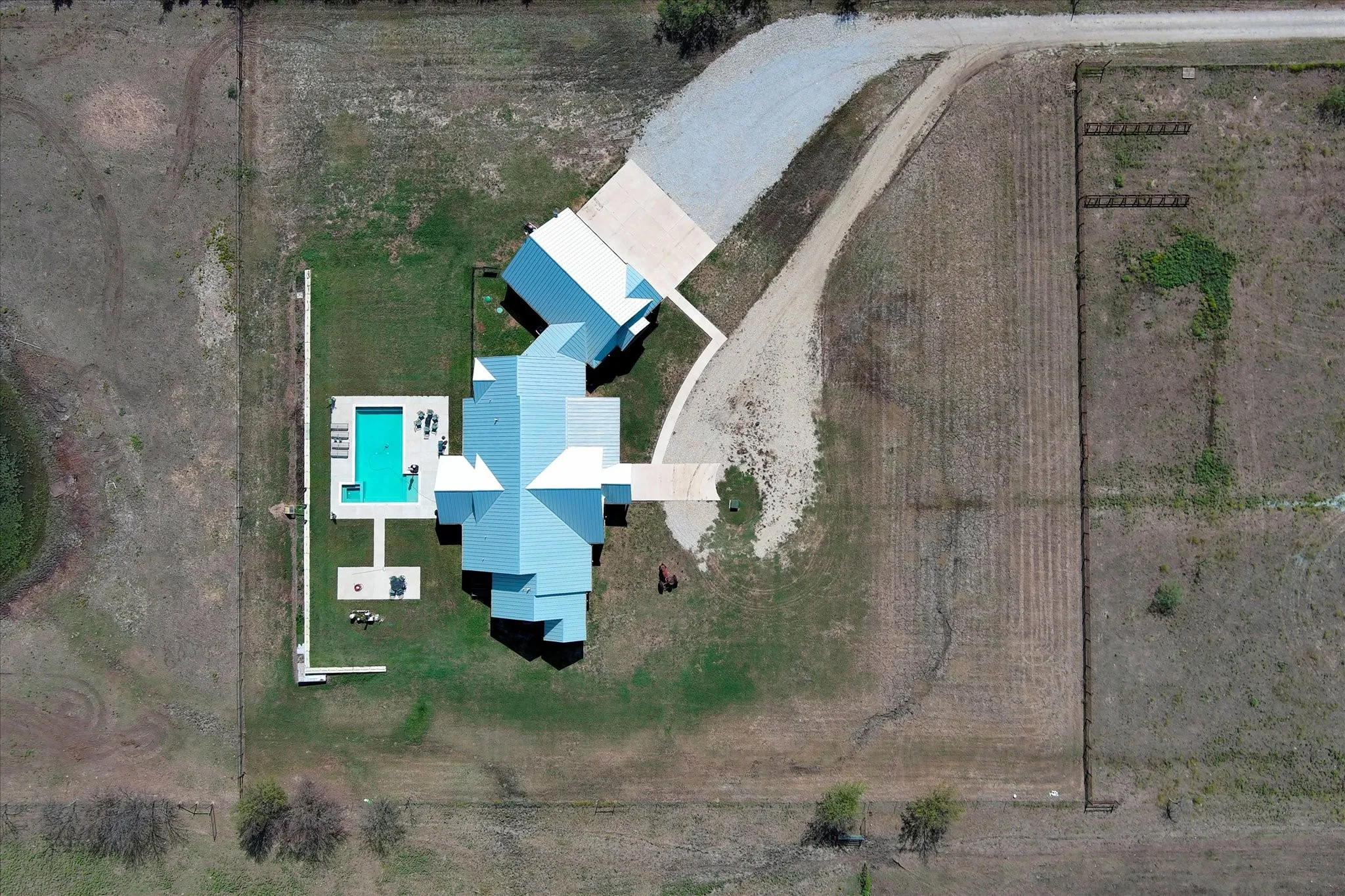 Aerial perspective of property featuring a metal roof, rectangular swimming pool, concrete patio, gravel driveway, and fenced perimeter