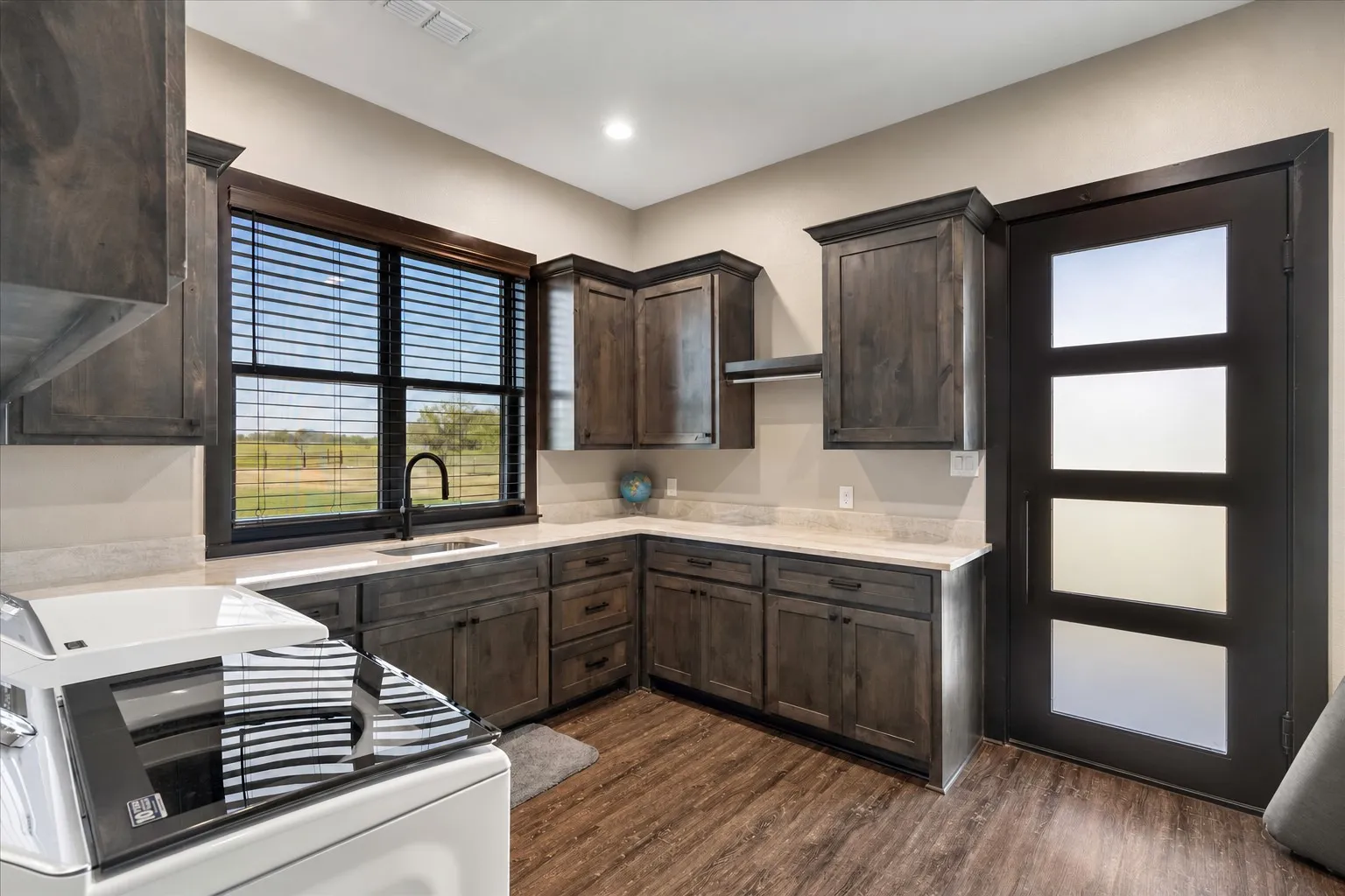 Custom dark-stained cabinetry with matte black hardware, light-toned stone-finish countertops, undermount sink with a matte black faucet, wood-finish flooring, and a modern exterior door featuring frosted glass panels