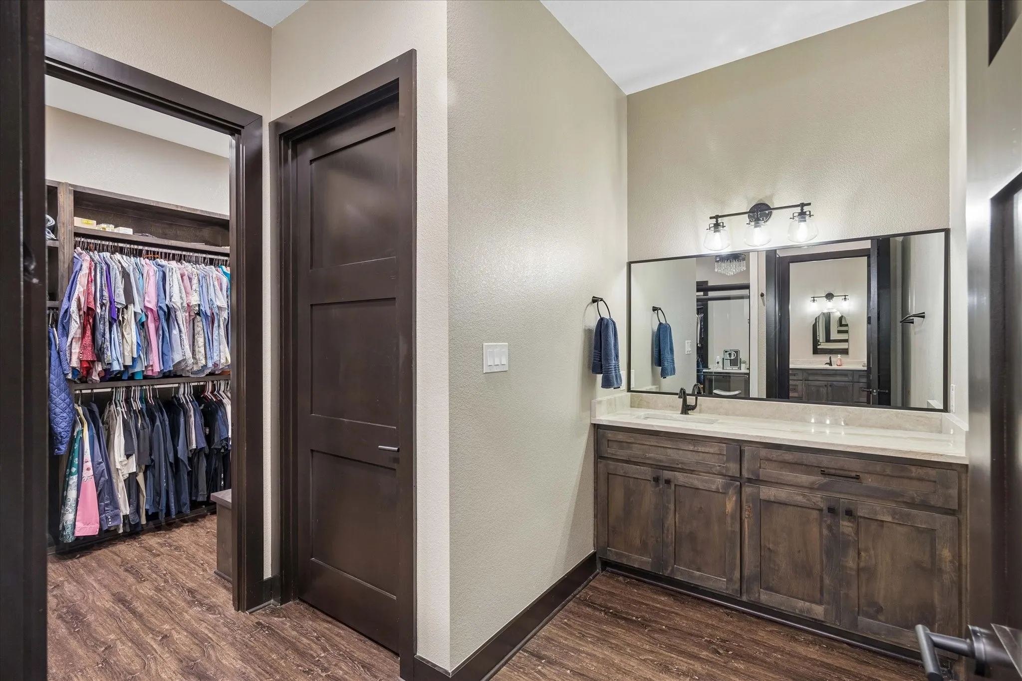 Bathroom featuring dual-sink vanity with stone countertop, dark wood-finish cabinetry, wood-finish flooring, and walk-in closet with built-in shelving