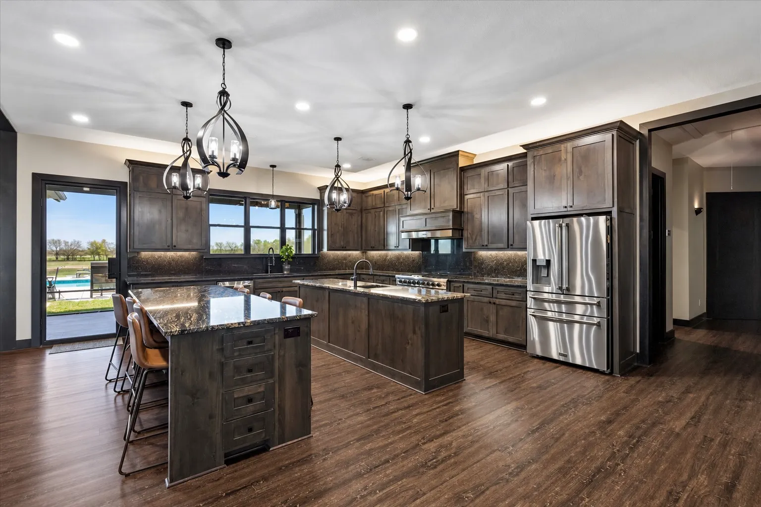 Kitchen featuring dark-finish cabinetry, two islands with stone countertops, stainless steel appliances, wood-finish flooring, and pendant lighting