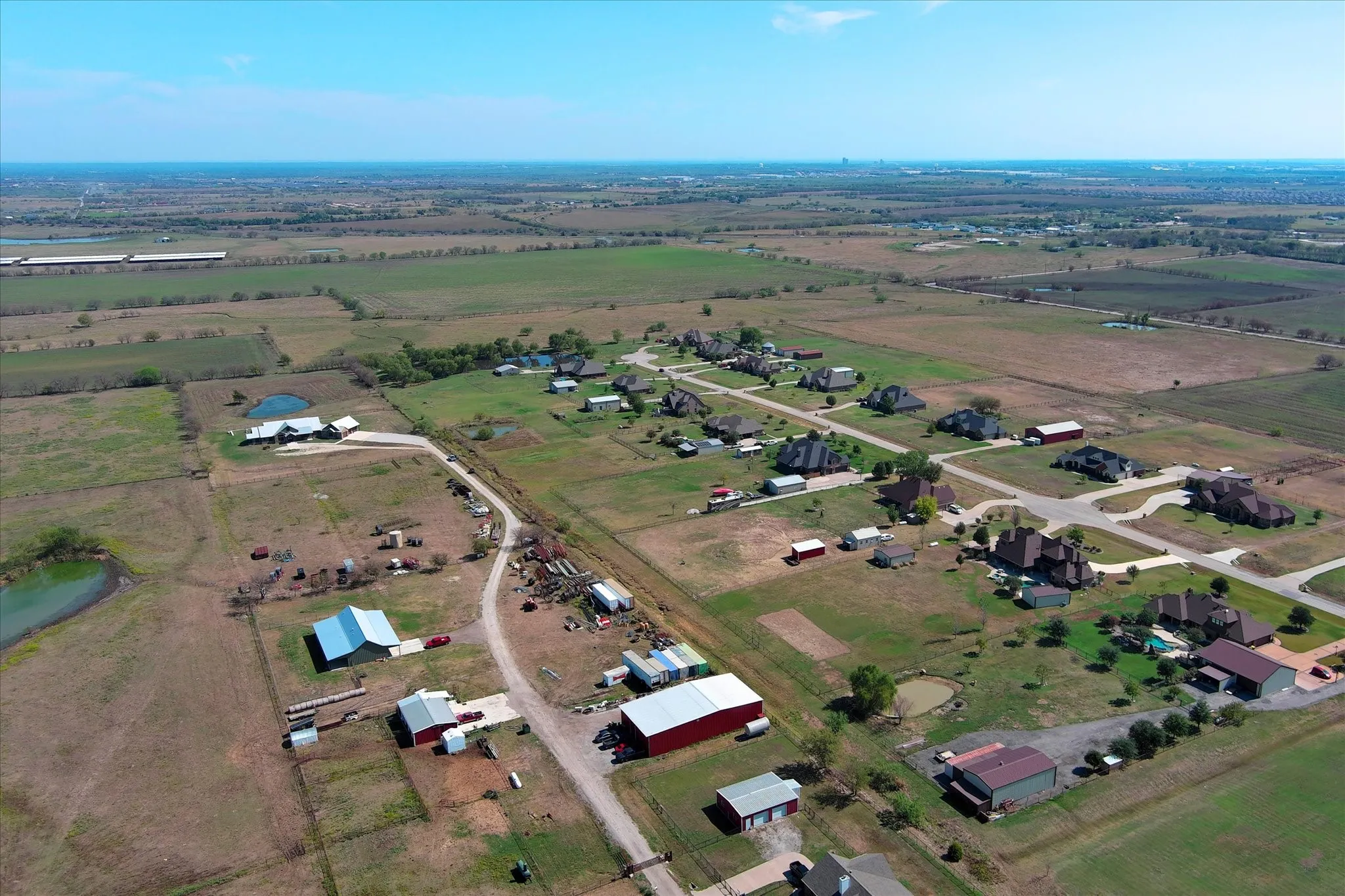Aerial perspective of expansive acreage featuring multiple metal-roofed outbuildings, fenced pastures, and paved driveway access