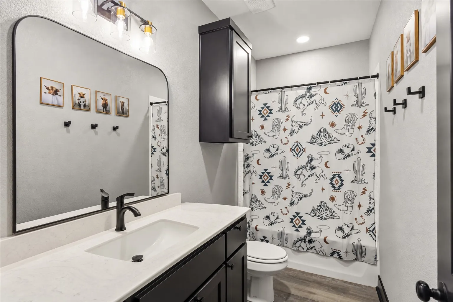 Bathroom featuring a vanity with white stone-style countertop and undermount sink, dark cabinetry, arched wall-mounted mirror, three-light fixture with clear glass shades, and wood-finish flooring
