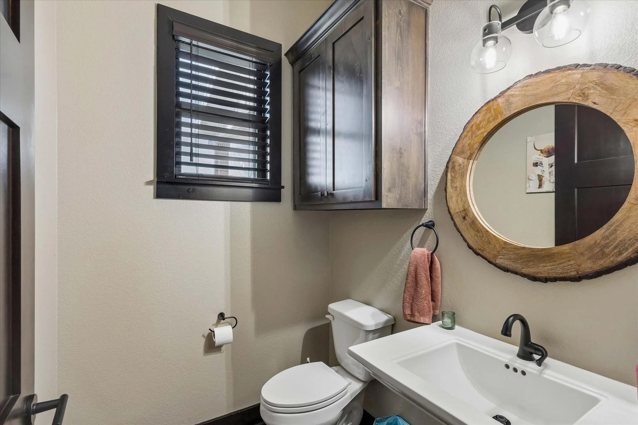 Restroom featuring a wall-mounted wood-finish cabinet, natural edge circular mirror, white basin with matte black faucet, and two-light vanity fixture