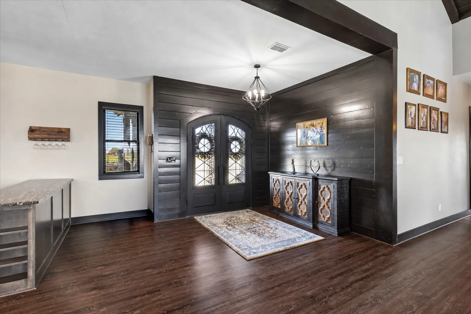 Double arched front doors with decorative glass inserts, black horizontal wood-plank wall detailing, exposed ceiling beams, wood-finish flooring, and a modern geometric chandelier