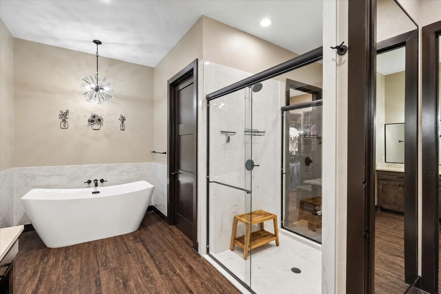Freestanding soaking tub with wall-mounted matte black fixtures, glass-enclosed walk-in shower with tile surround, wood-finish flooring, and statement starburst pendant lighting