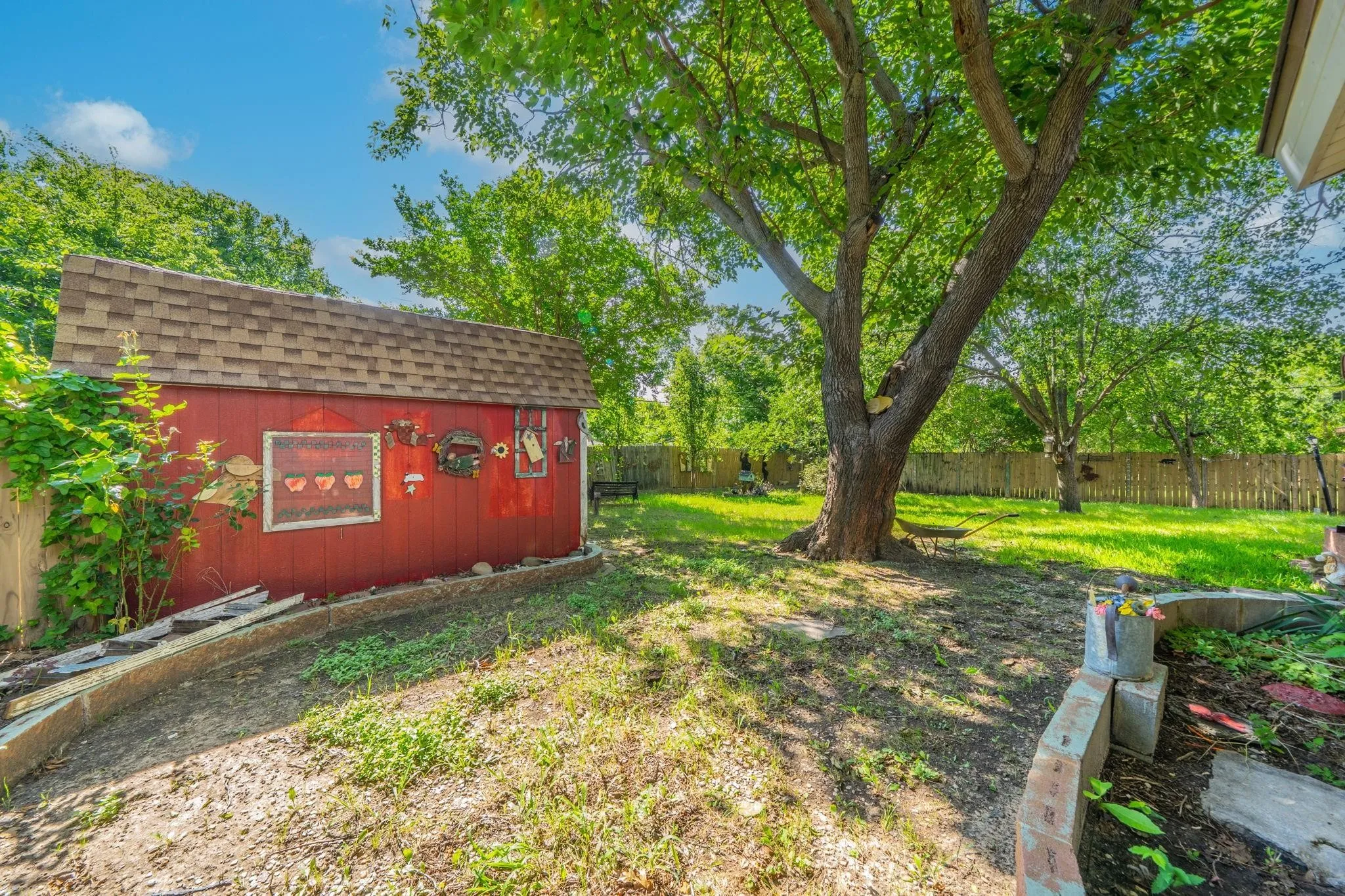 Backyard featuring a red storage shed with a shingled roof, mature tree, grass lawn, wood fencing, and tiered stone garden beds