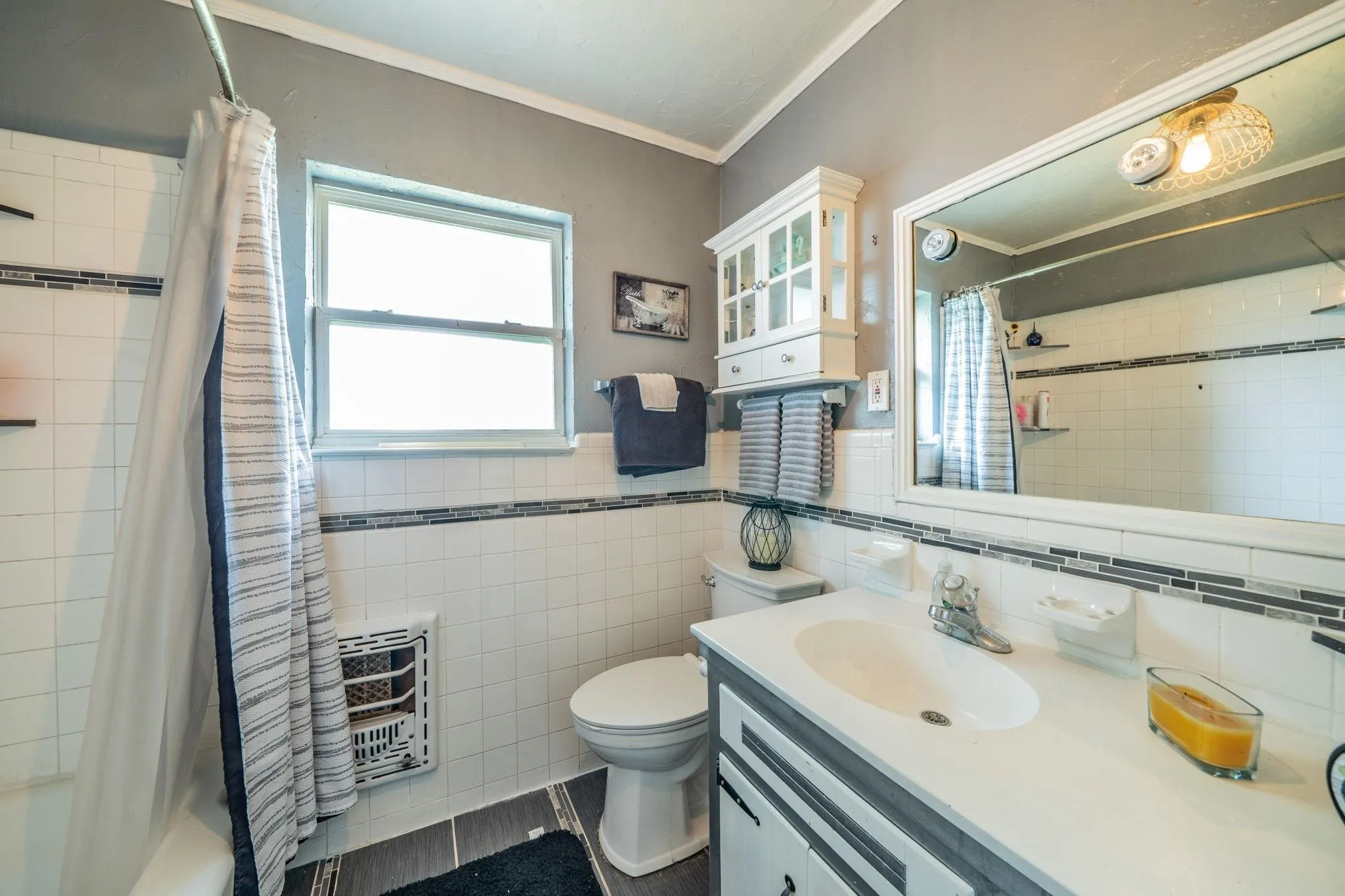 Bathroom featuring a vanity with a white countertop and oval basin, wall-mounted white cabinetry, a large rectangular mirror, and white tile wainscoting with decorative mosaic border accents