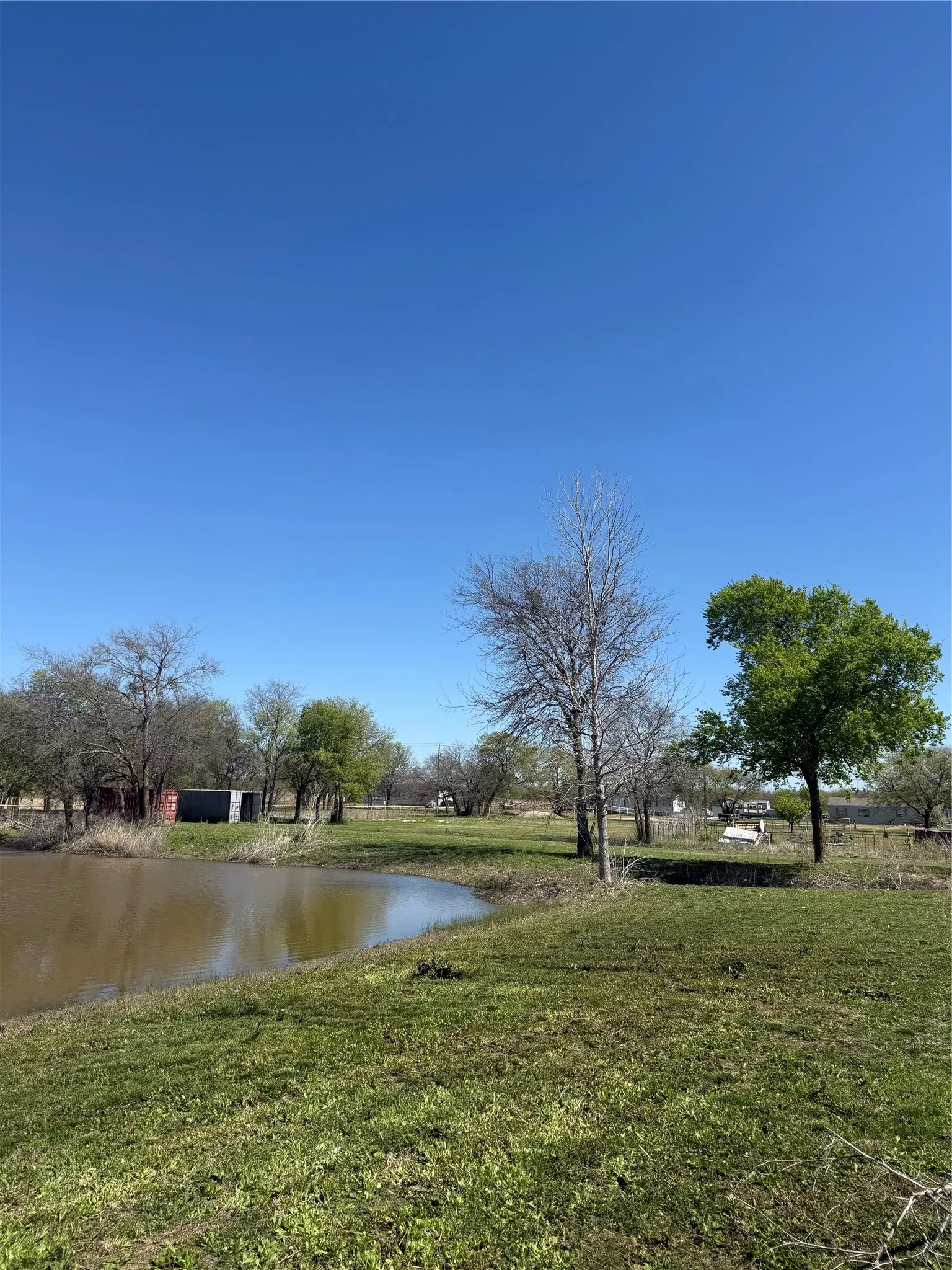 Expansive property featuring a curved pond, lush green grass, mature trees, a red barn, and a metal utility structure