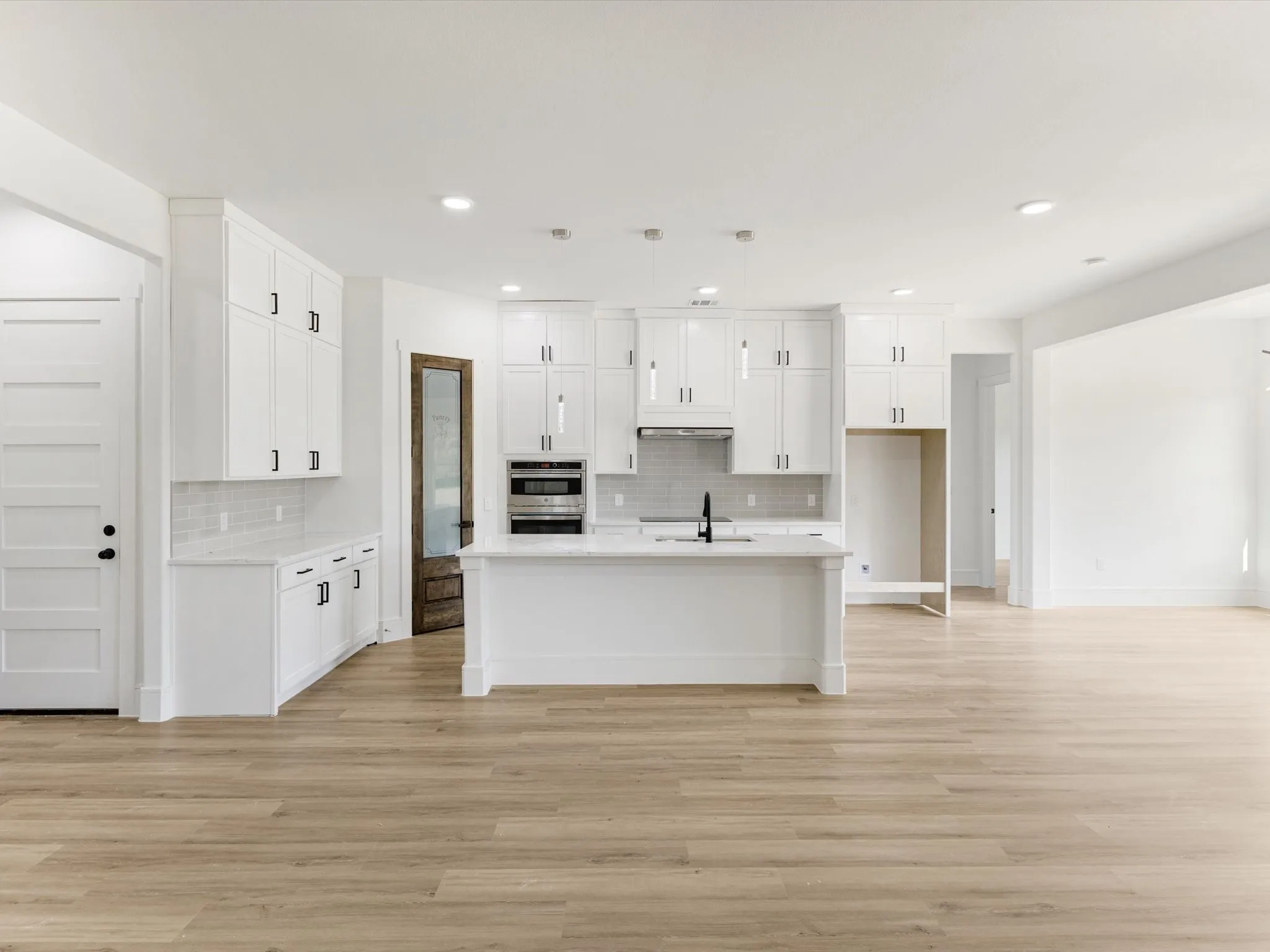 Kitchen featuring white ceiling-height cabinetry, a central island with a stone-finish countertop, wood-finish flooring, built-in stainless steel wall oven, and a pantry with a wood-toned door