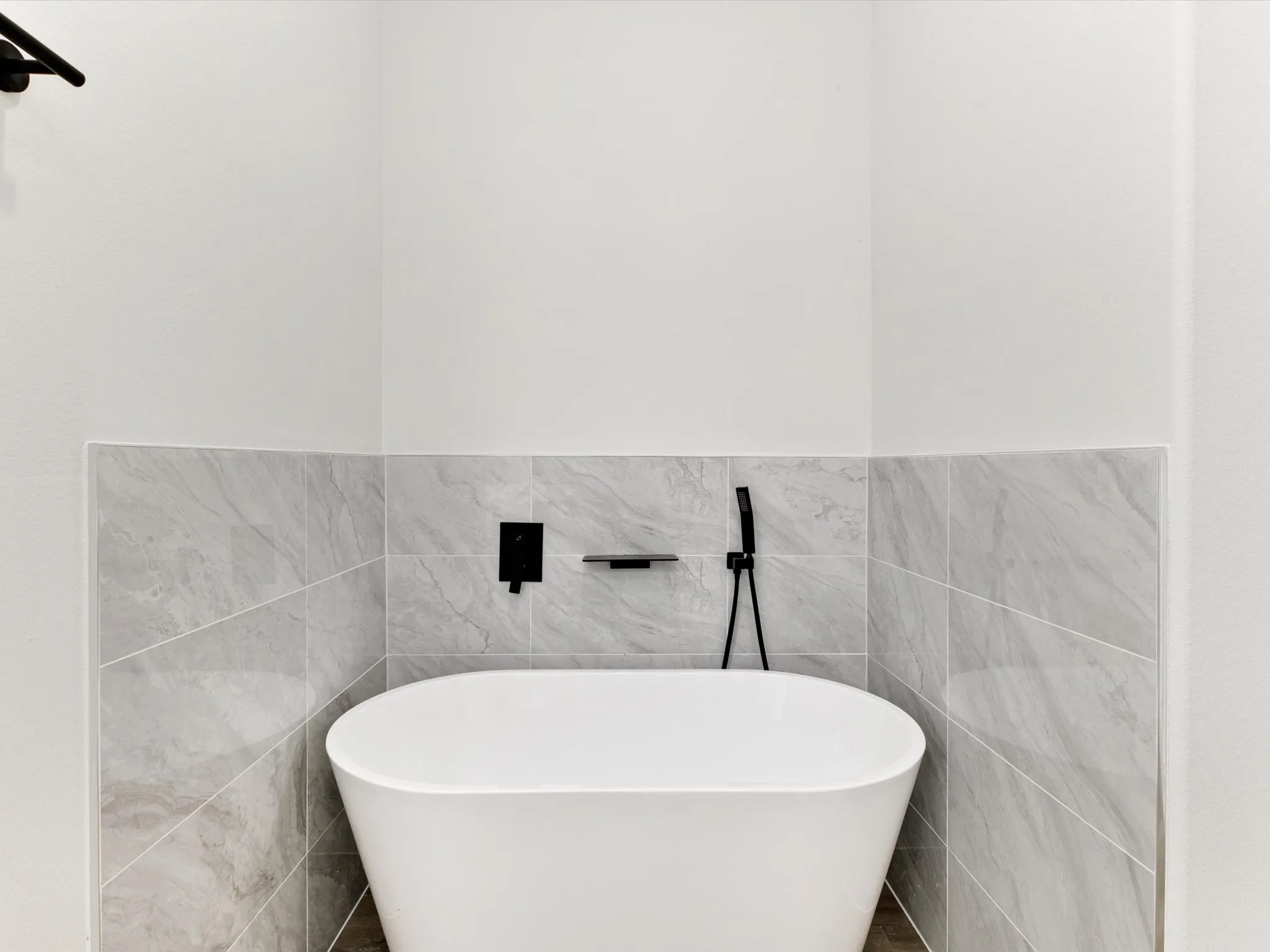 Freestanding white soaking tub with matte black wall-mounted faucet, handheld shower wand, built-in soap niche, and grey stone-finish tile surround
