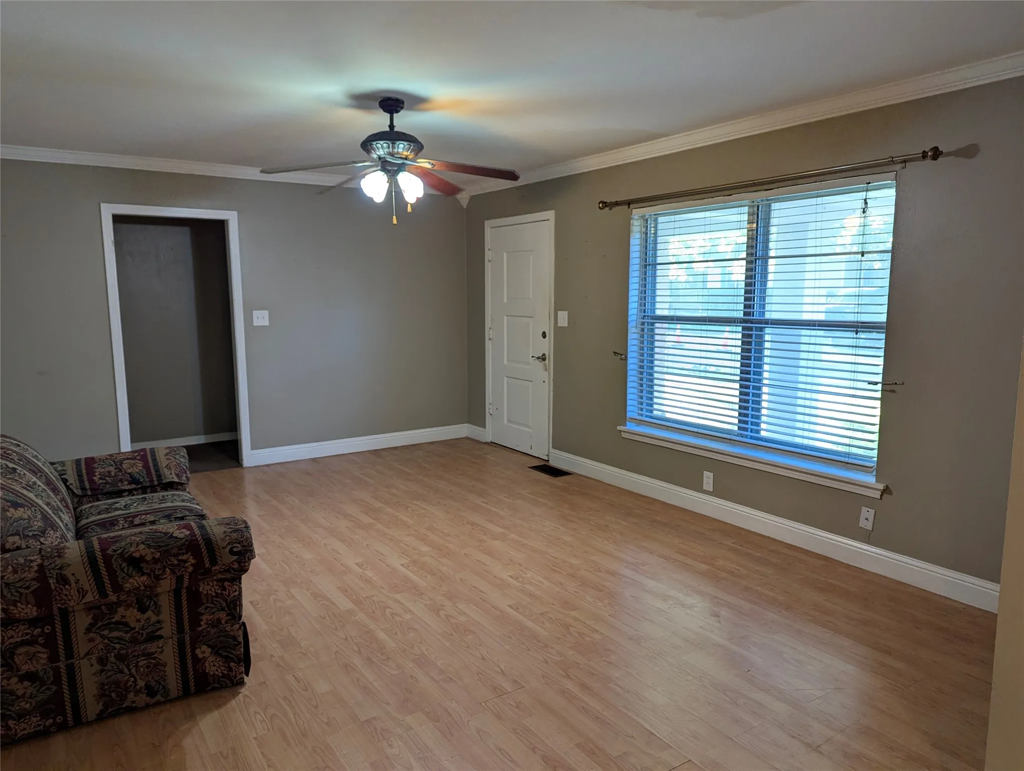 Spacious living room featuring wood-look flooring, neutral wall colors, crown molding, and a ceiling fan with lighting. Large front window welcomes natural light, creating a comfortable space ready for your personal touches!