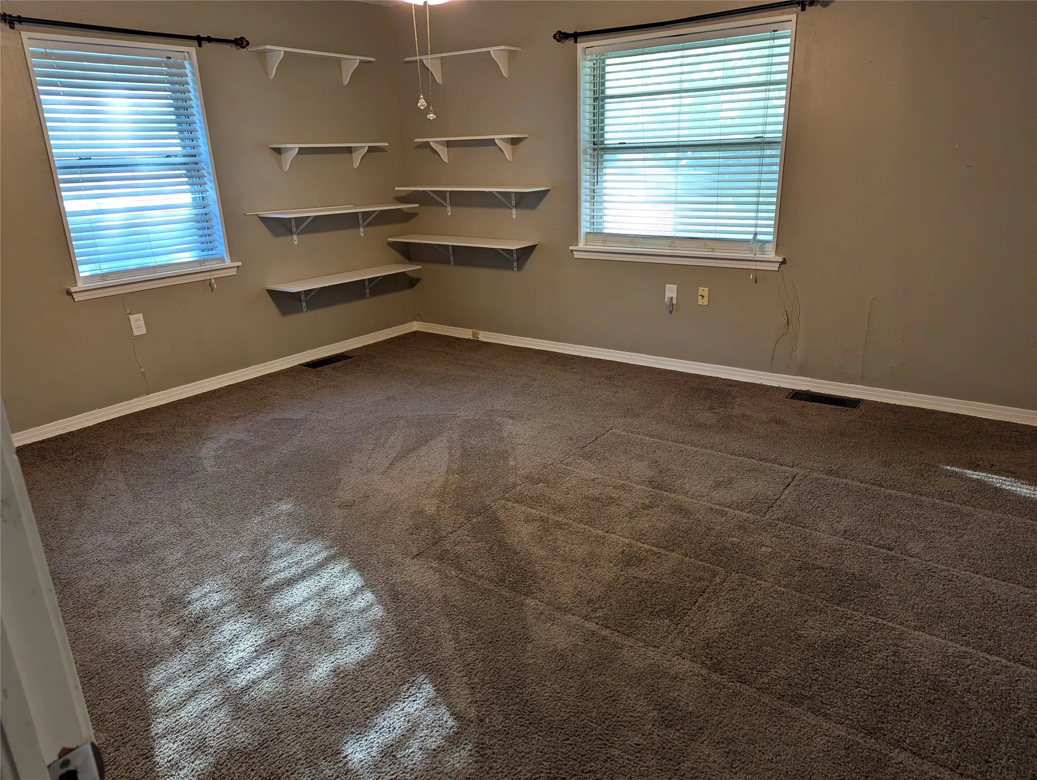 Bedroom #3 – View 1: Spacious secondary bedroom located in the southeast corner of the home, featuring two windows overlooking the front yard, carpeted flooring, and built-in corner shelving.