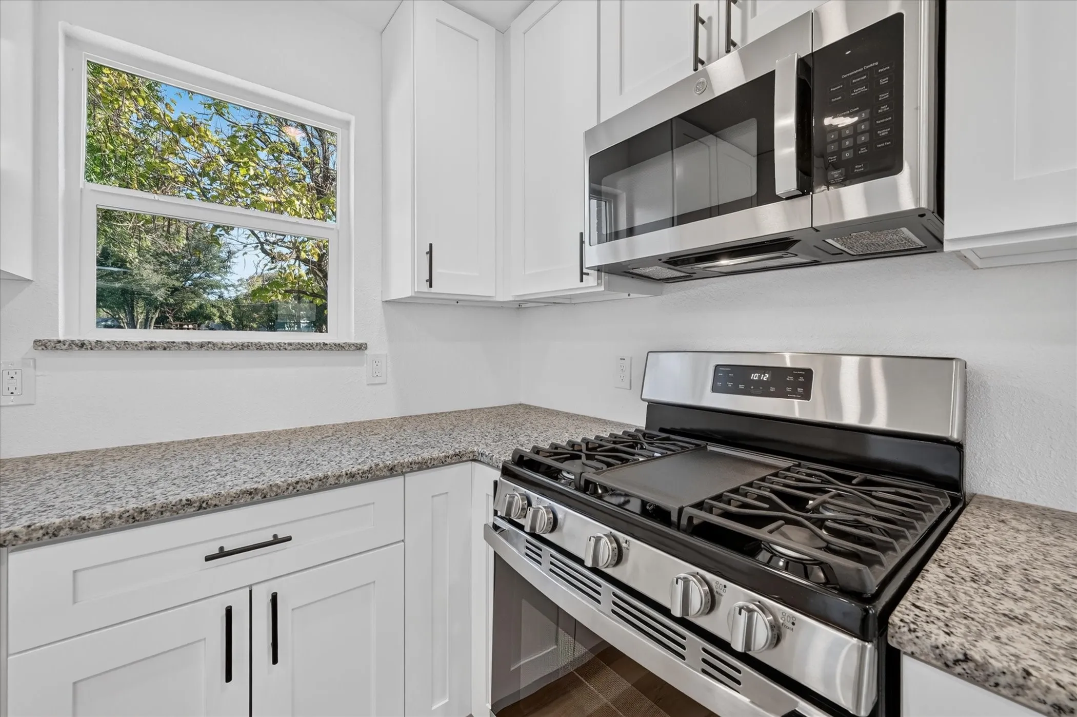 White shaker-style cabinetry, speckled stone countertops, stainless steel gas range with a griddle, over-the-range microwave, and a single-hung window with a stone sill