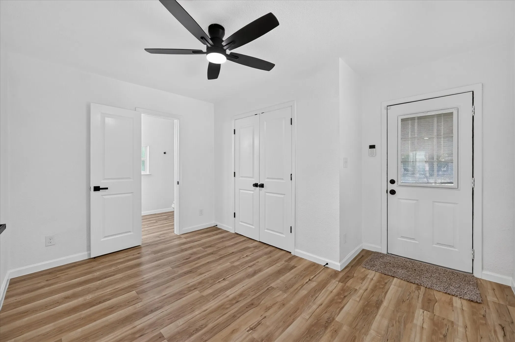 Wood-finish flooring, matte black hardware, modern ceiling fan with integrated lighting, dual-panel closet doors, and exterior door with inset window