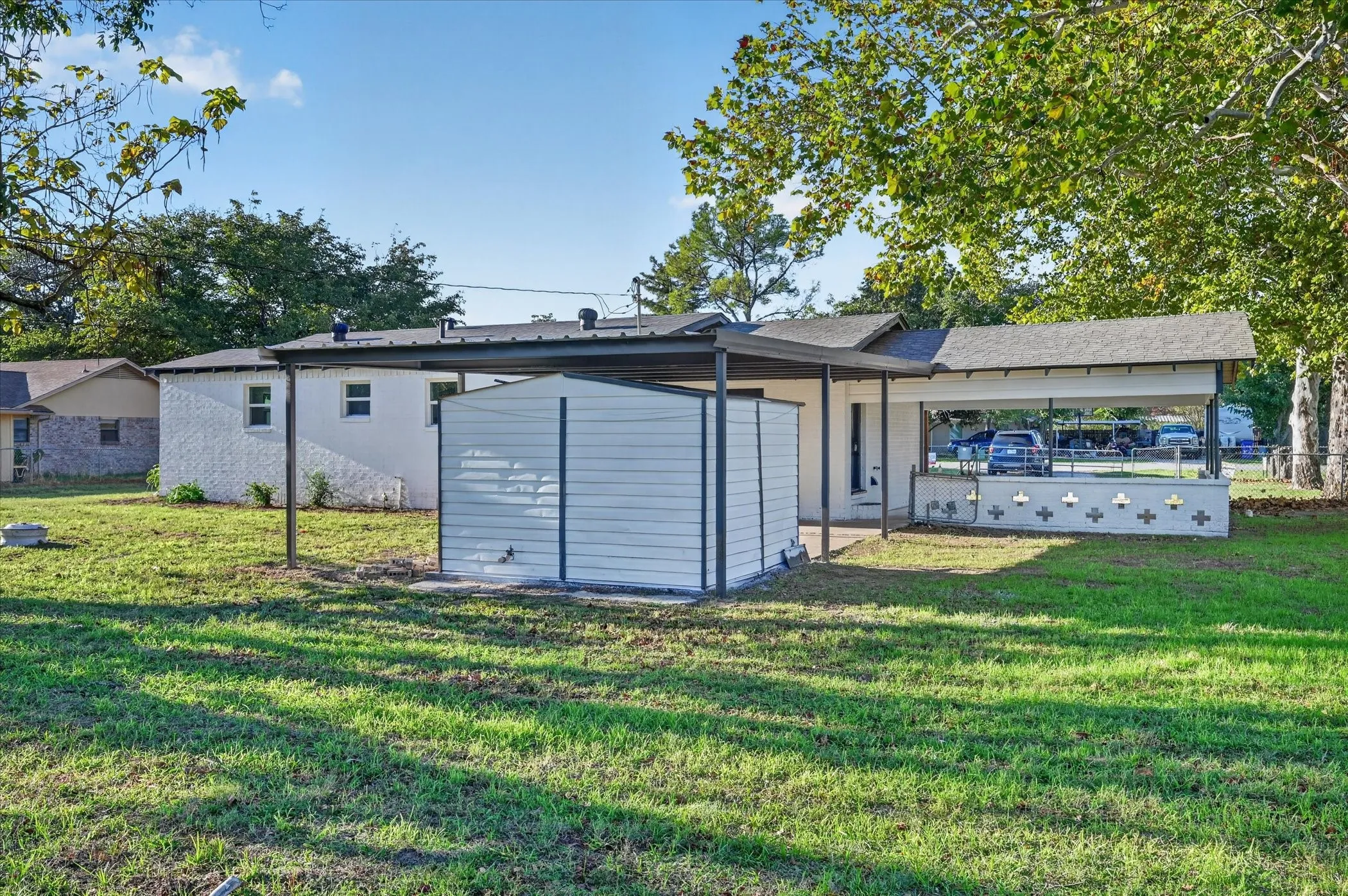 White brick exterior with a metal-roofed carport, decorative concrete block screening, and an attached storage unit situated on a grass lot