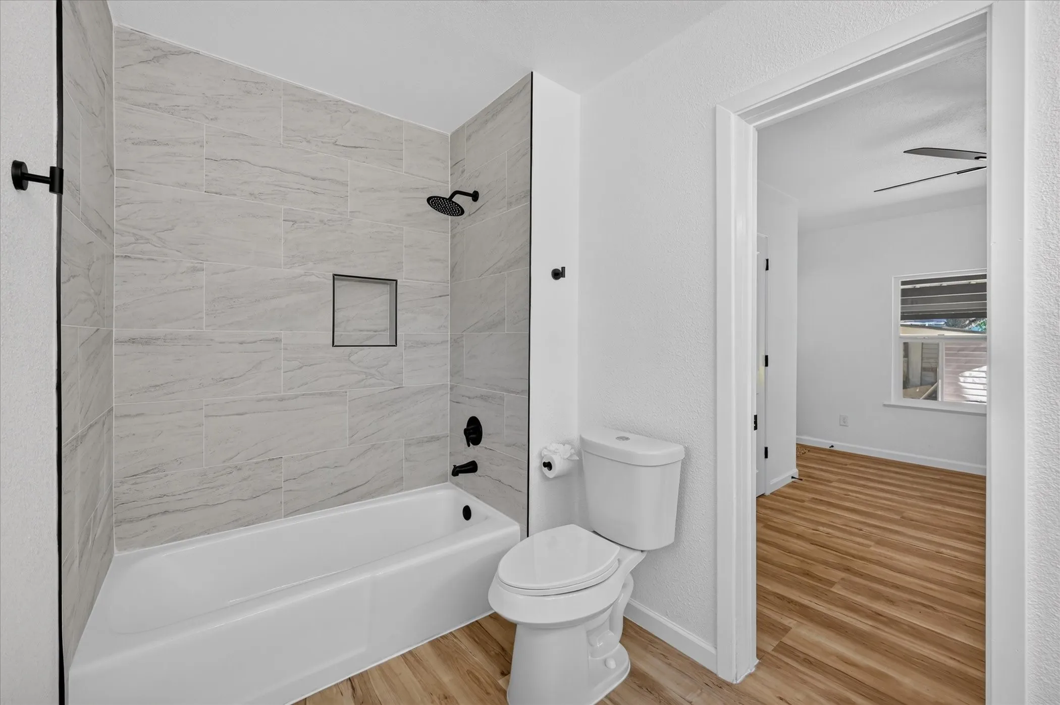 Bathroom featuring neutral-toned tile shower surround with a built-in niche, matte black fixtures, and wood-finish flooring