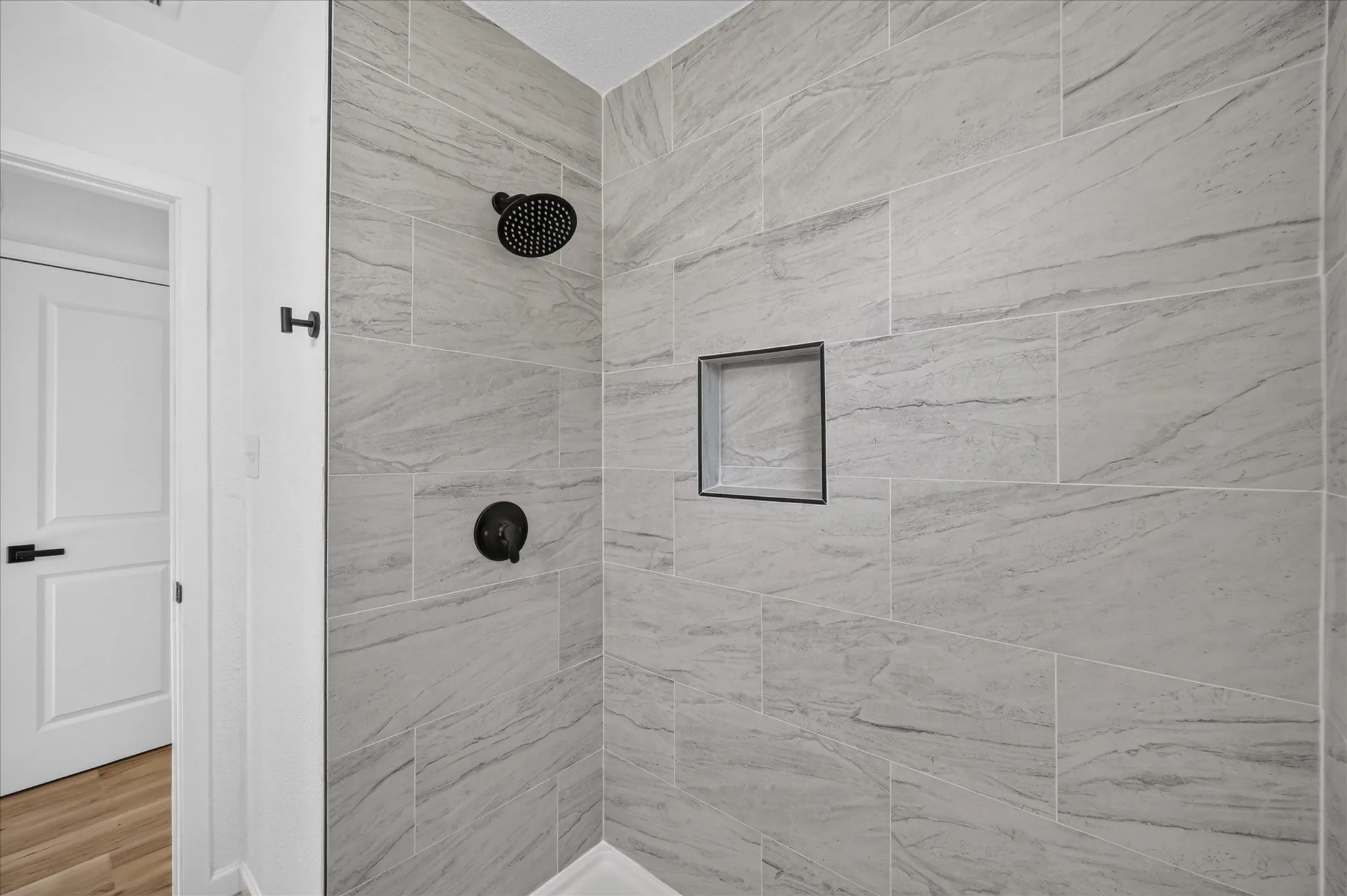 Walk-in shower featuring large-format grey stone-patterned tile, matte black rainfall showerhead, integrated tile niche, and matching matte black faucet controls