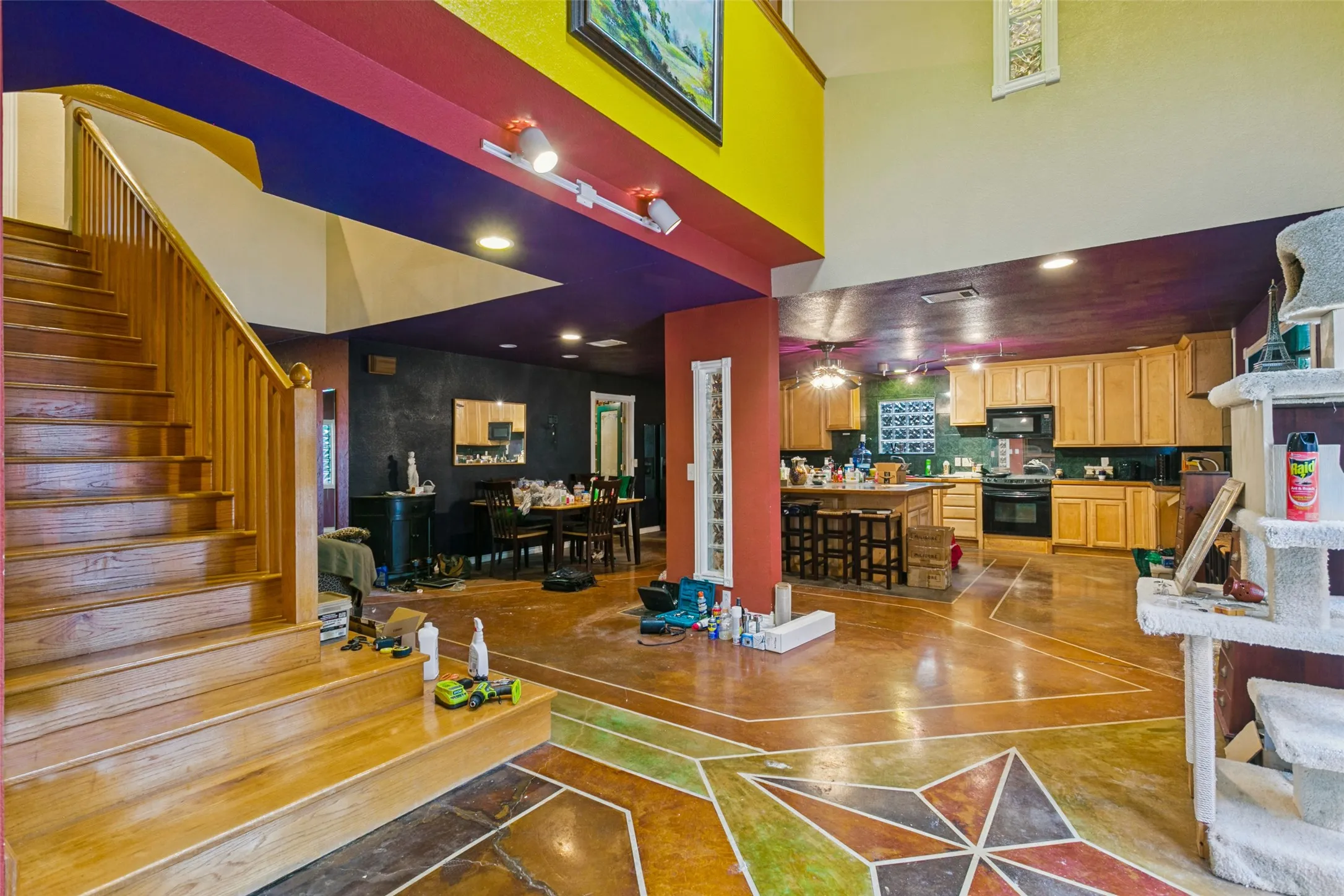 Open-concept interior featuring high ceilings, stained concrete flooring with decorative geometric inlays, a wooden staircase with traditional railings, kitchen cabinetry, and track lighting