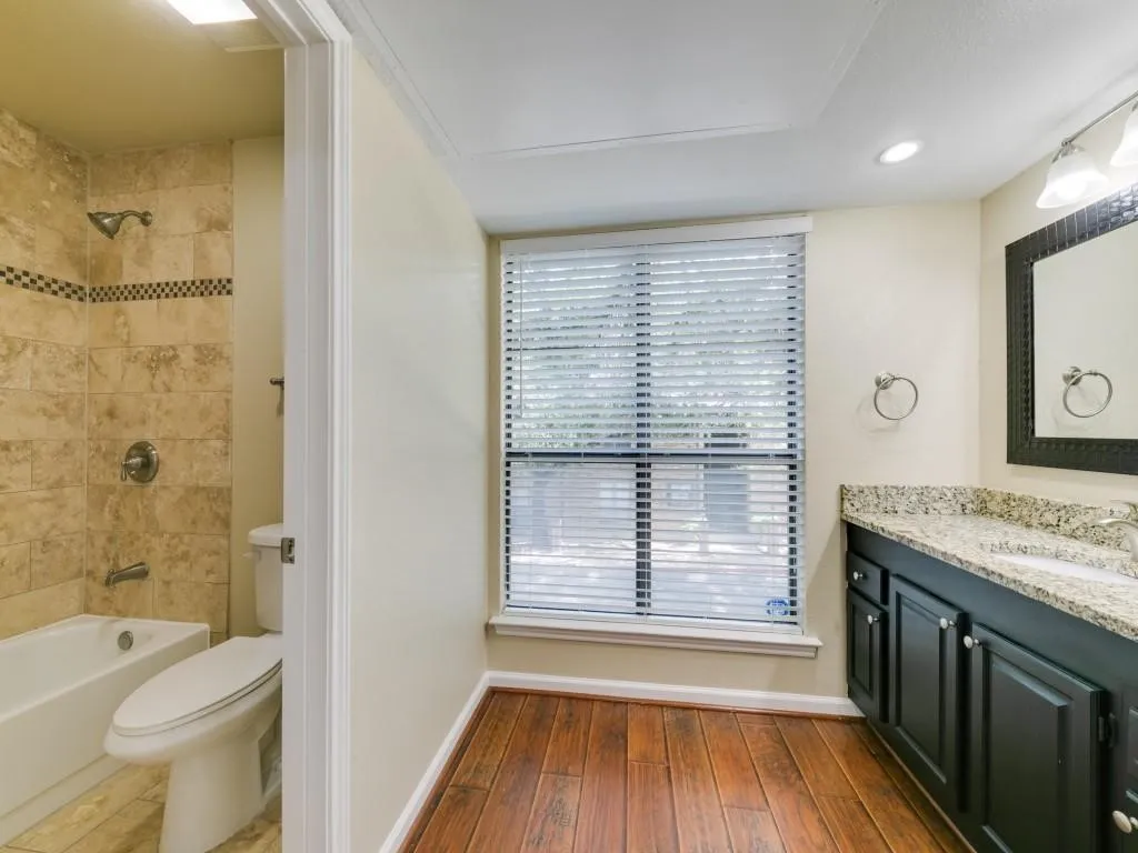 Bathroom featuring wood-finish flooring, dark-finish cabinetry with a stone-composite countertop, and a shower-tub combination with tile surround and a decorative mosaic inlay