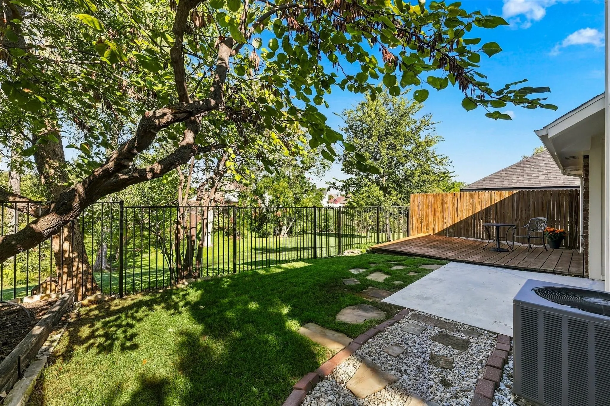 Backyard featuring a wood deck, stone-bordered gravel pathway, manicured lawn, and metal perimeter fencing