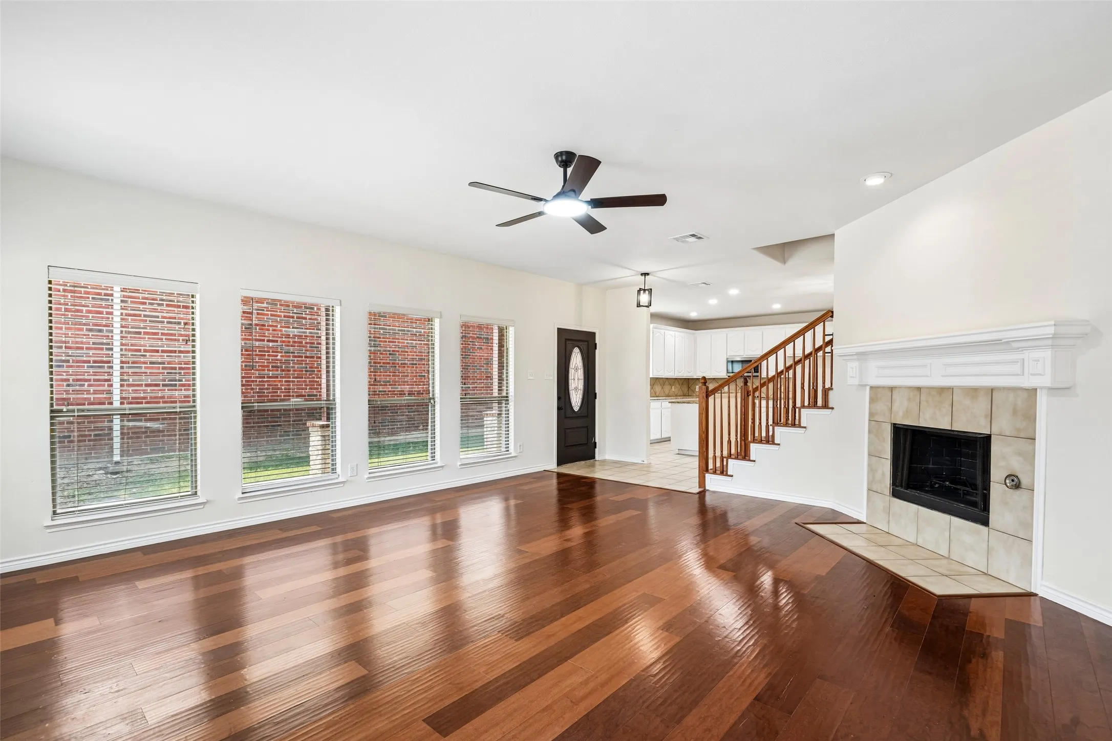 Open-concept interior featuring wood-finish flooring, a fireplace with a ceramic tile surround, four large windows, and a staircase with a wooden balustrade