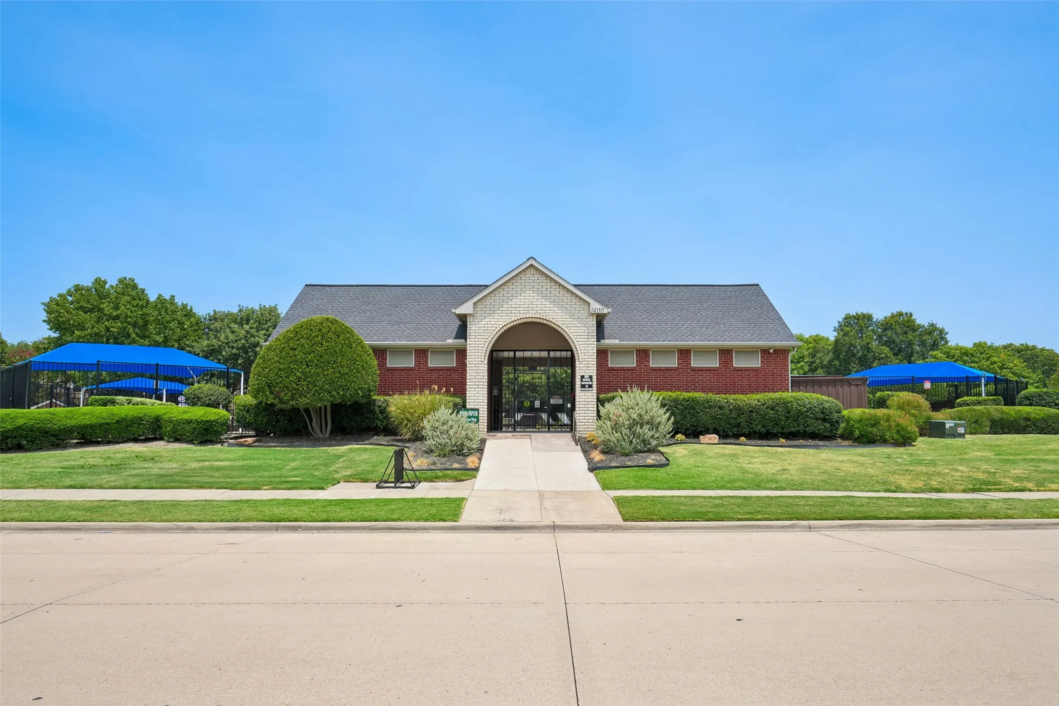 Brick clubhouse exterior featuring an arched entrance, shingled roof, manicured landscaping, and blue shade structures
