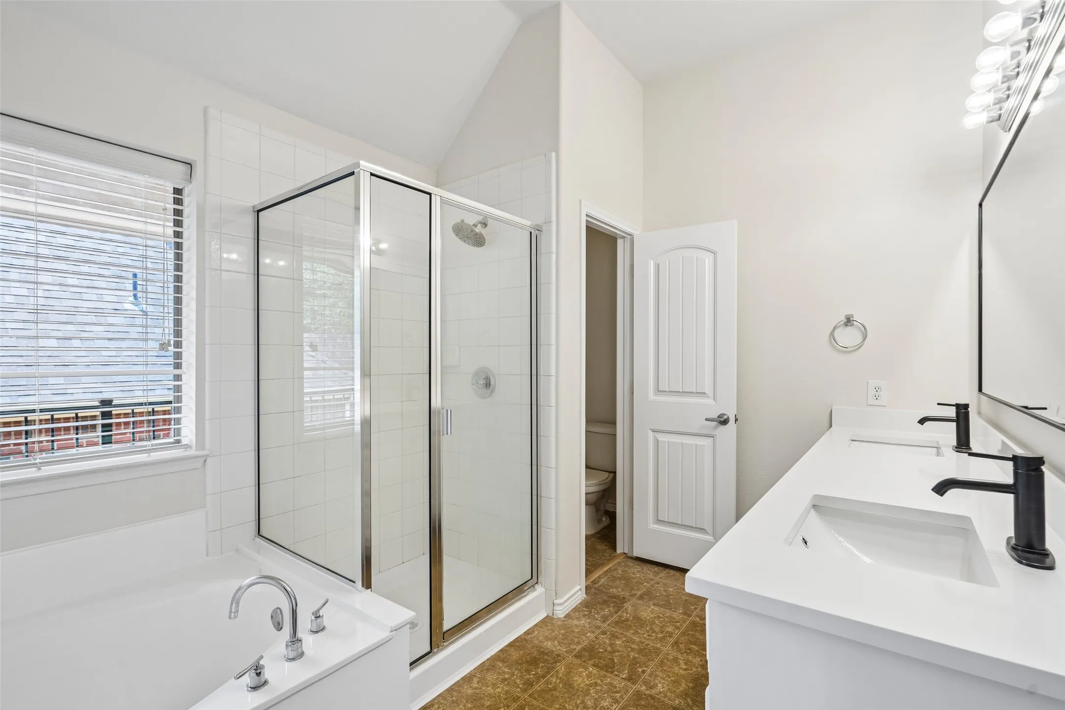 Bathroom featuring a dual-sink vanity with white countertops, matte black fixtures, a glass-enclosed shower, a deep soaking tub, and tile flooring