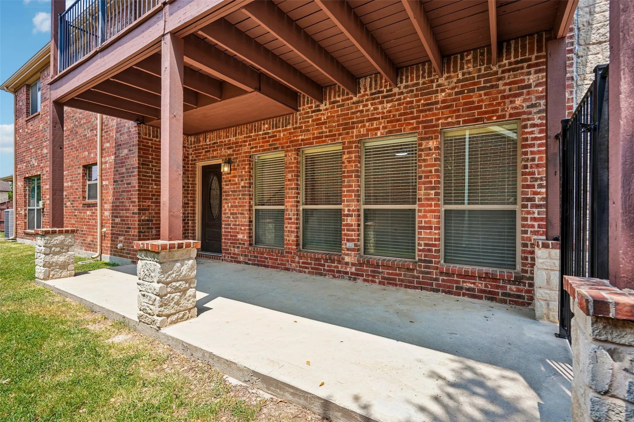Covered concrete patio featuring brick exterior walls, stone-based support columns, and an overhead balcony with metal railings