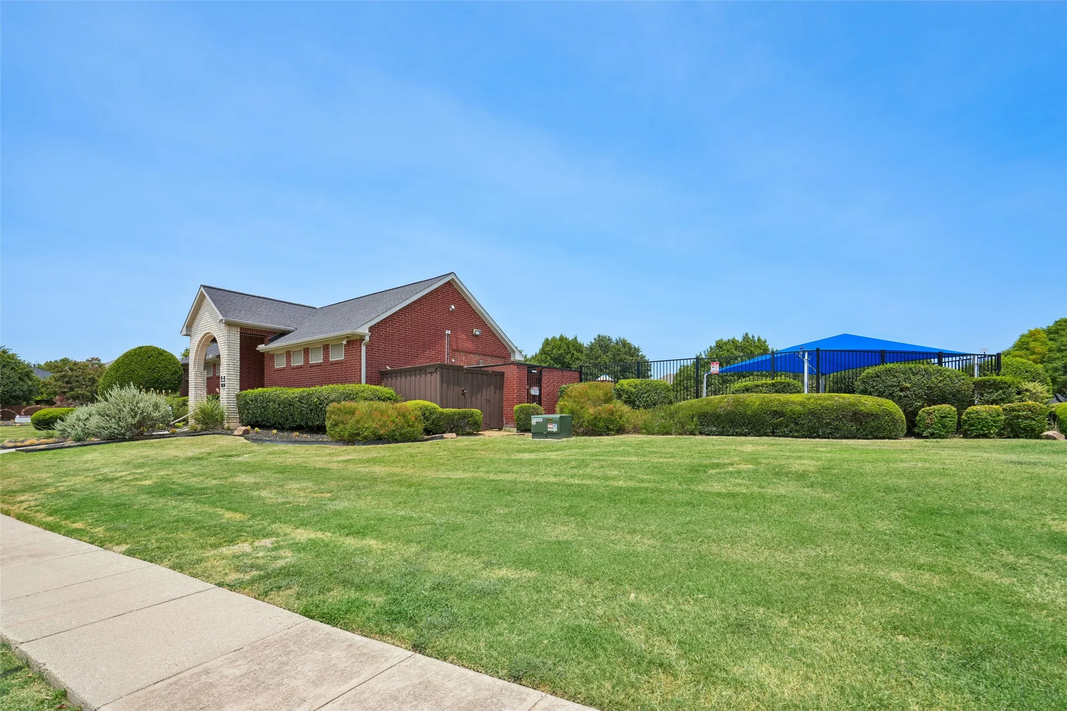 Red brick exterior featuring a covered entryway, manicured lawn, landscaping, and a fenced area with shade structures