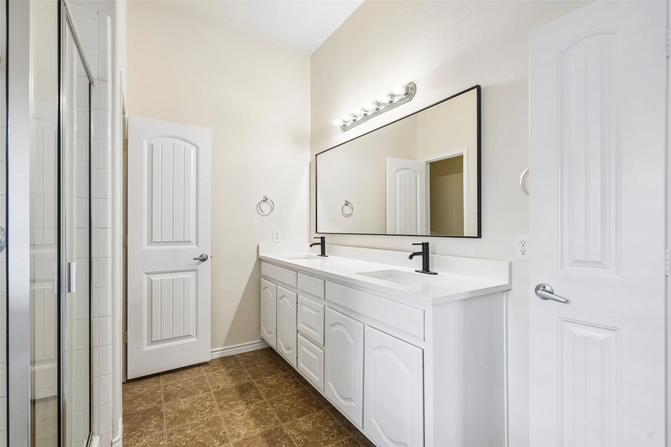 Dual-sink vanity with white countertop, matte black faucets, oversized rectangular mirror, and glass-enclosed shower stall