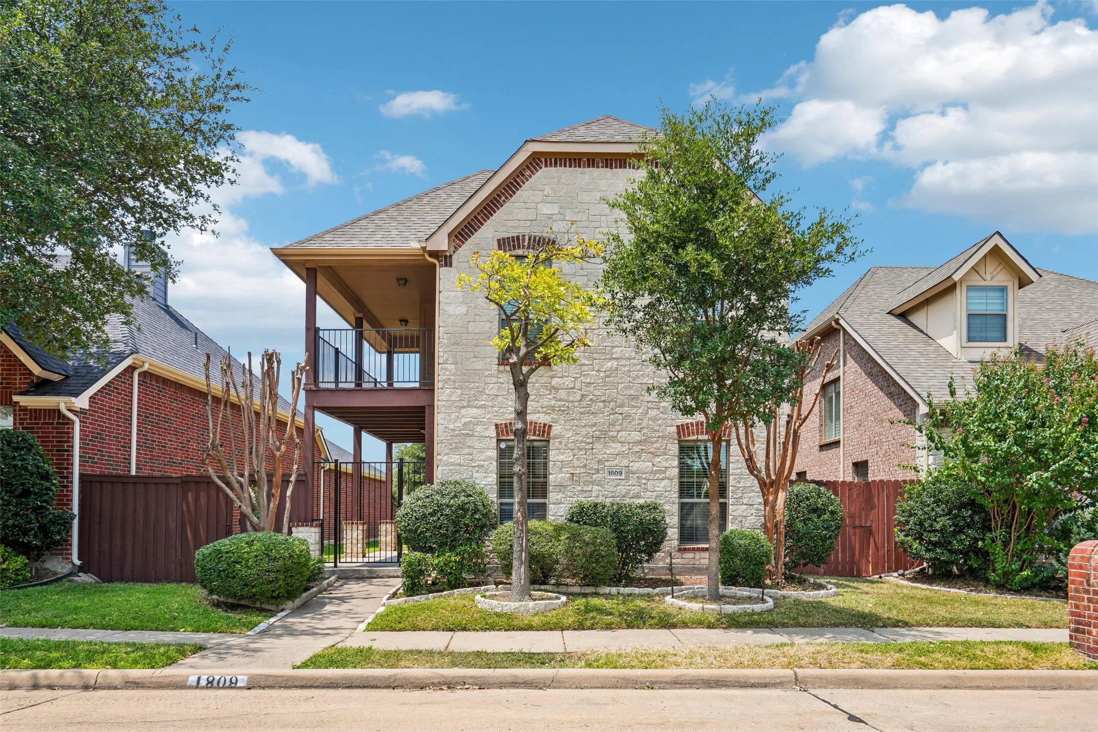 Two-story exterior featuring light-toned stone masonry, a covered second-story balcony with metal railing, gabled rooflines, and mature landscaping